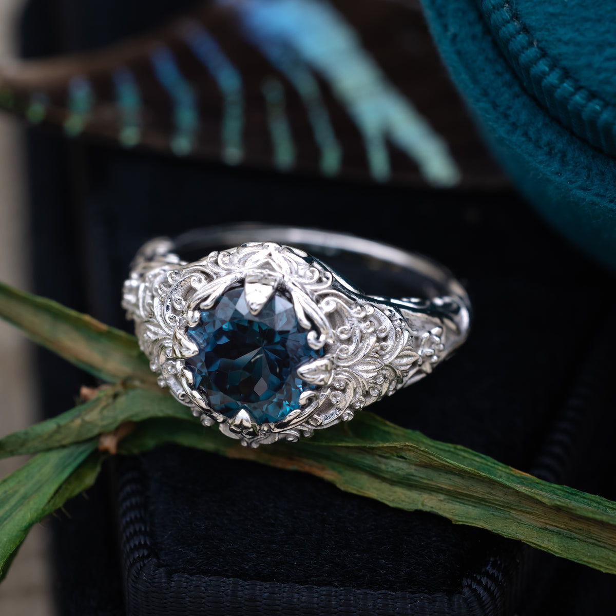 White gold ring with sculptural filigree and round alexandrite center stone on a dark blue cushions with greenery