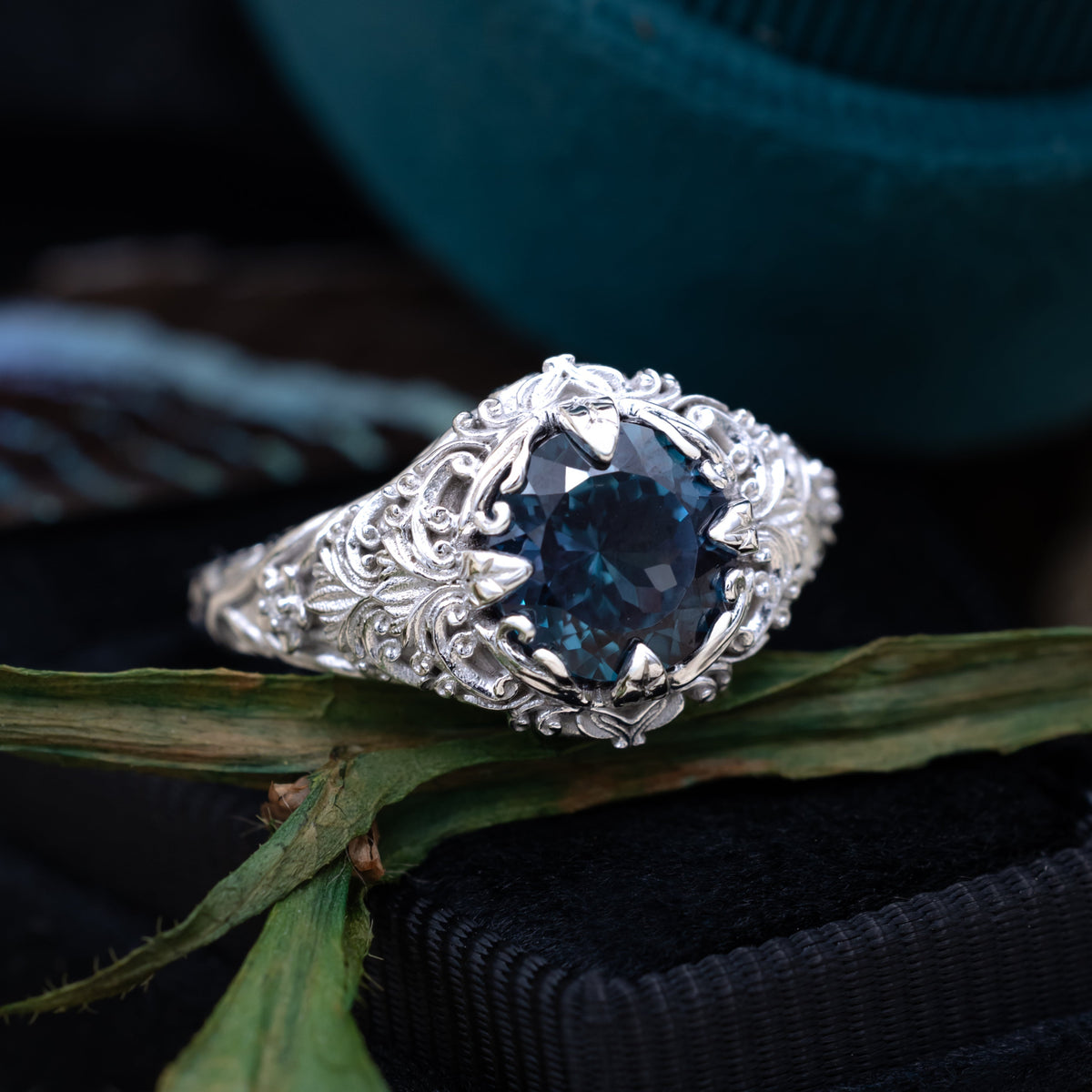 White gold ring with sculptural filigree and round alexandrite center stone on a dark blue cushions with greenery