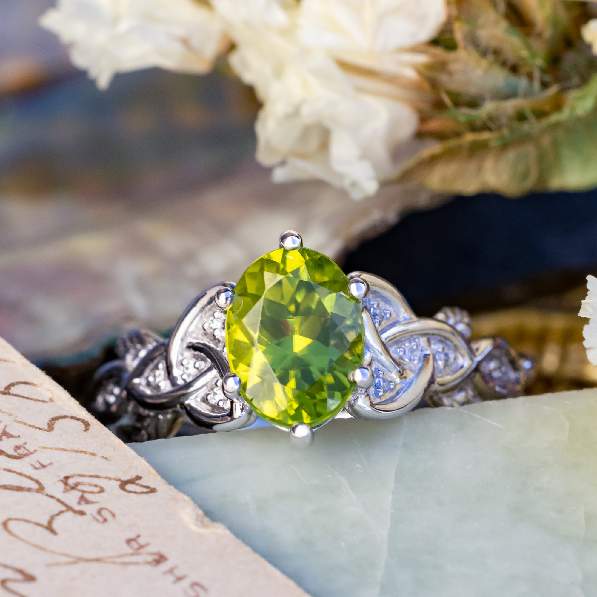 White gold ring with a green peridot on a textured surface with flowers in the background