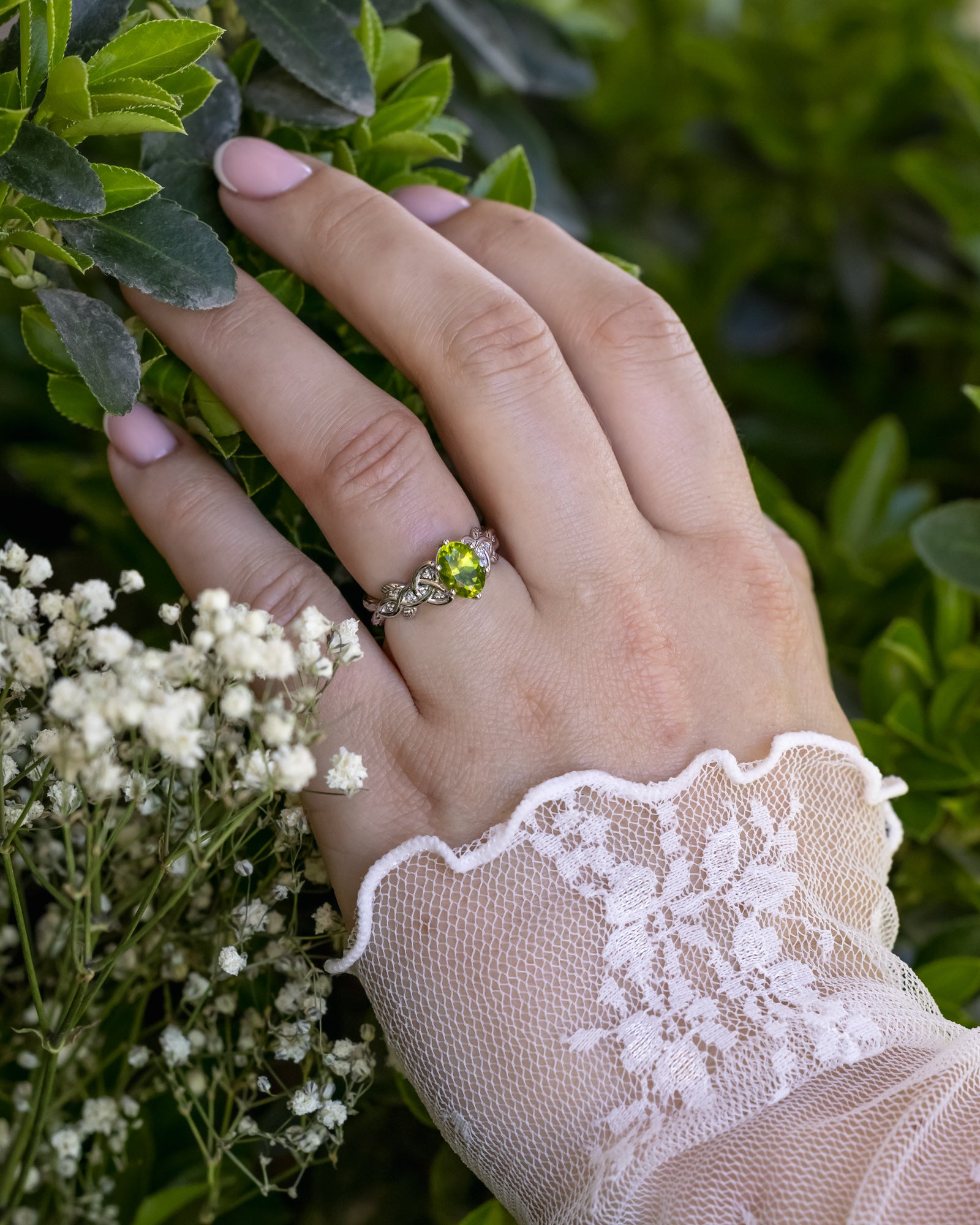 Hand wearing a ring with a green peridot, surrounded by greenery and white flowers.
