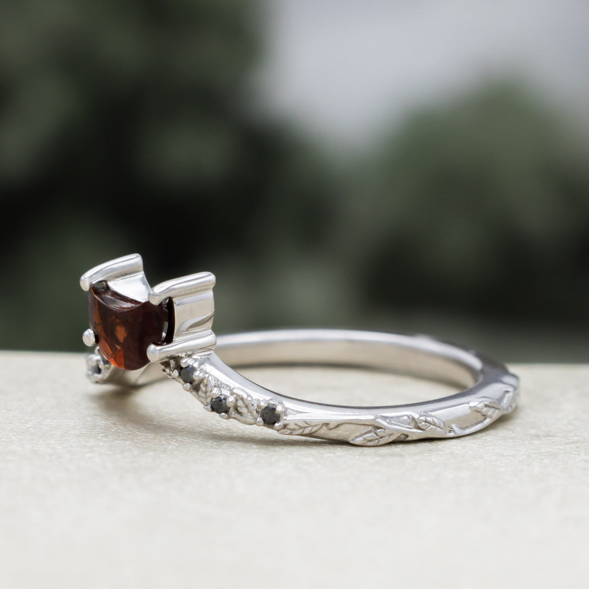 White gold crescent moon garnet tracer with vine band and black diamond accents against a blurred green background