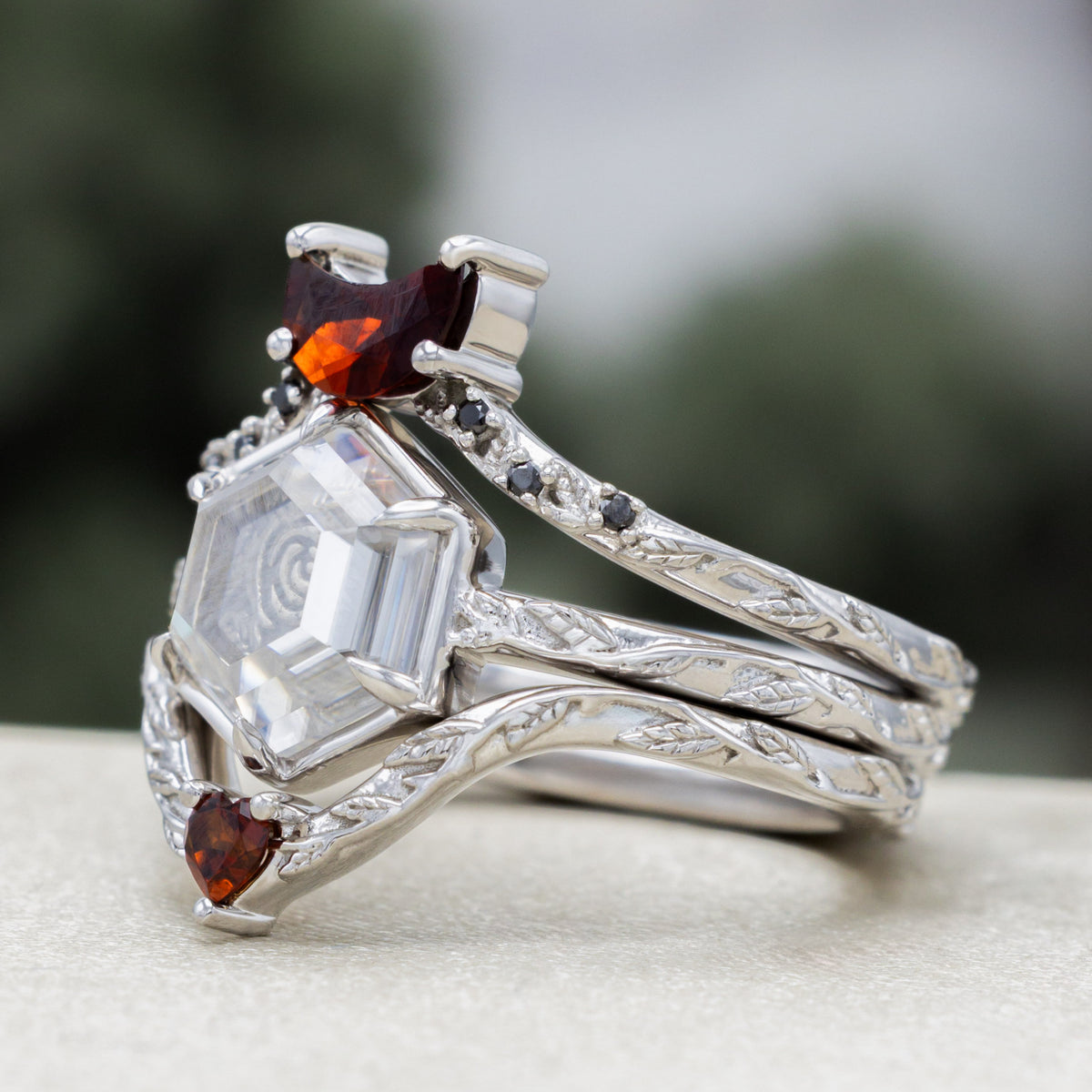 White gold engagement ring with portrait cut moissanite featuring rose design and matching garnet tracers on vine band against a blurred green background