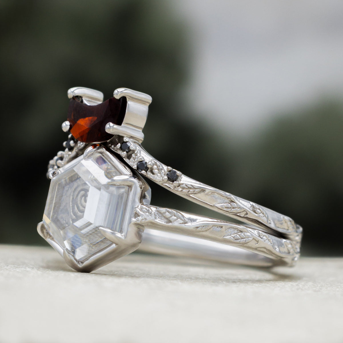 White gold engagement ring with portrait cut moissanite featuring rose design and crescent moon garnet tracer on vine band against a blurred green background