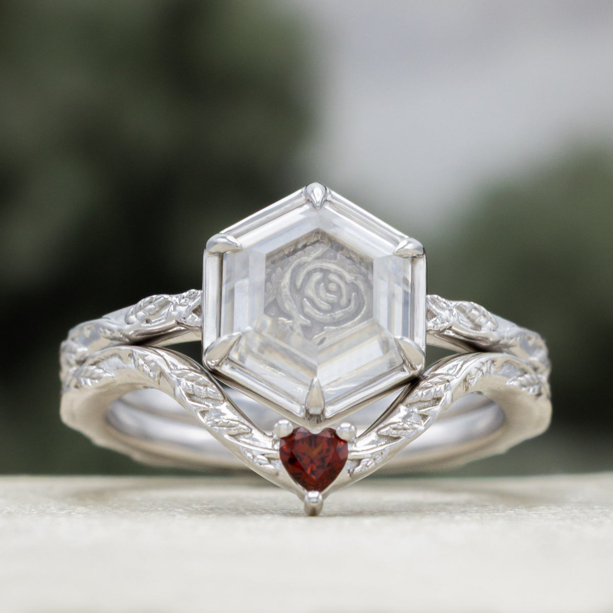 White gold engagement ring with portrait cut moissanite featuring rose design and heart garnet tracer on vine band against a blurred green background