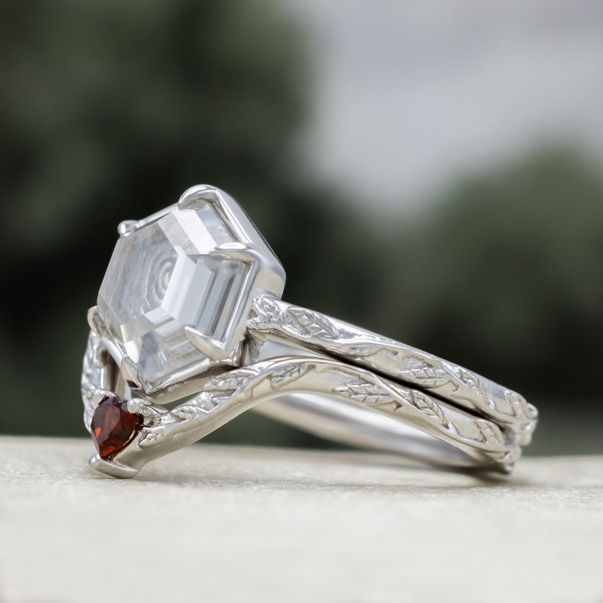White gold engagement ring with portrait cut moissanite featuring rose design and heart garnet tracer on vine band against a blurred green background