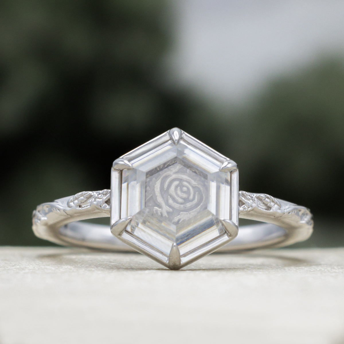 White gold engagement ring with portrait cut moissanite featuring rose design on vine band against a blurred green background