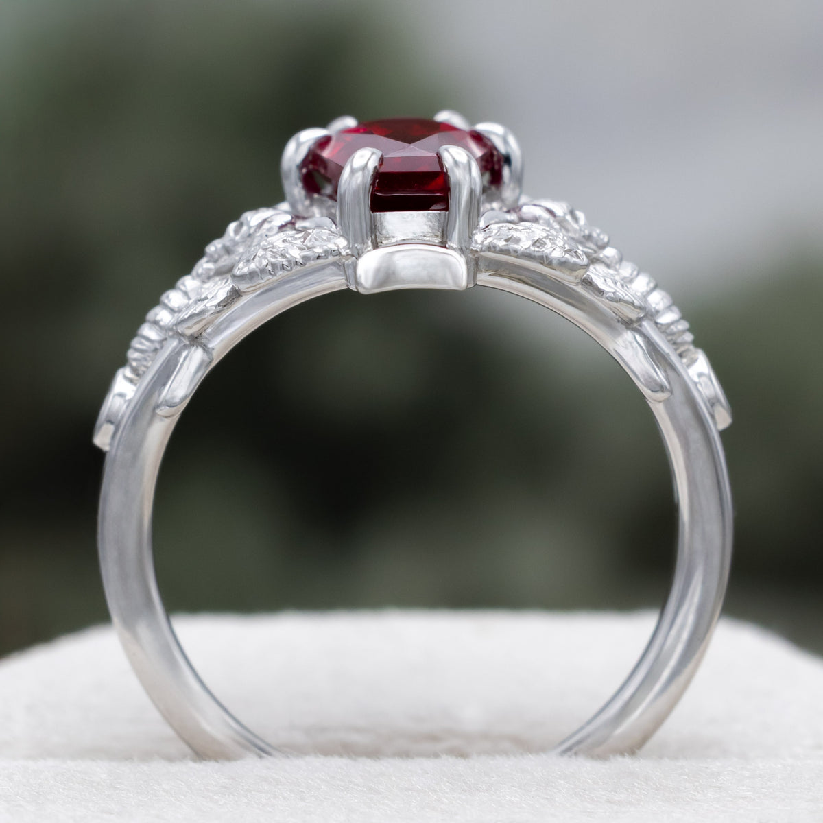 White gold engagement ring with deadhead moth filigree, coffin cut ruby center stone, and diamond accents against a blurred green background