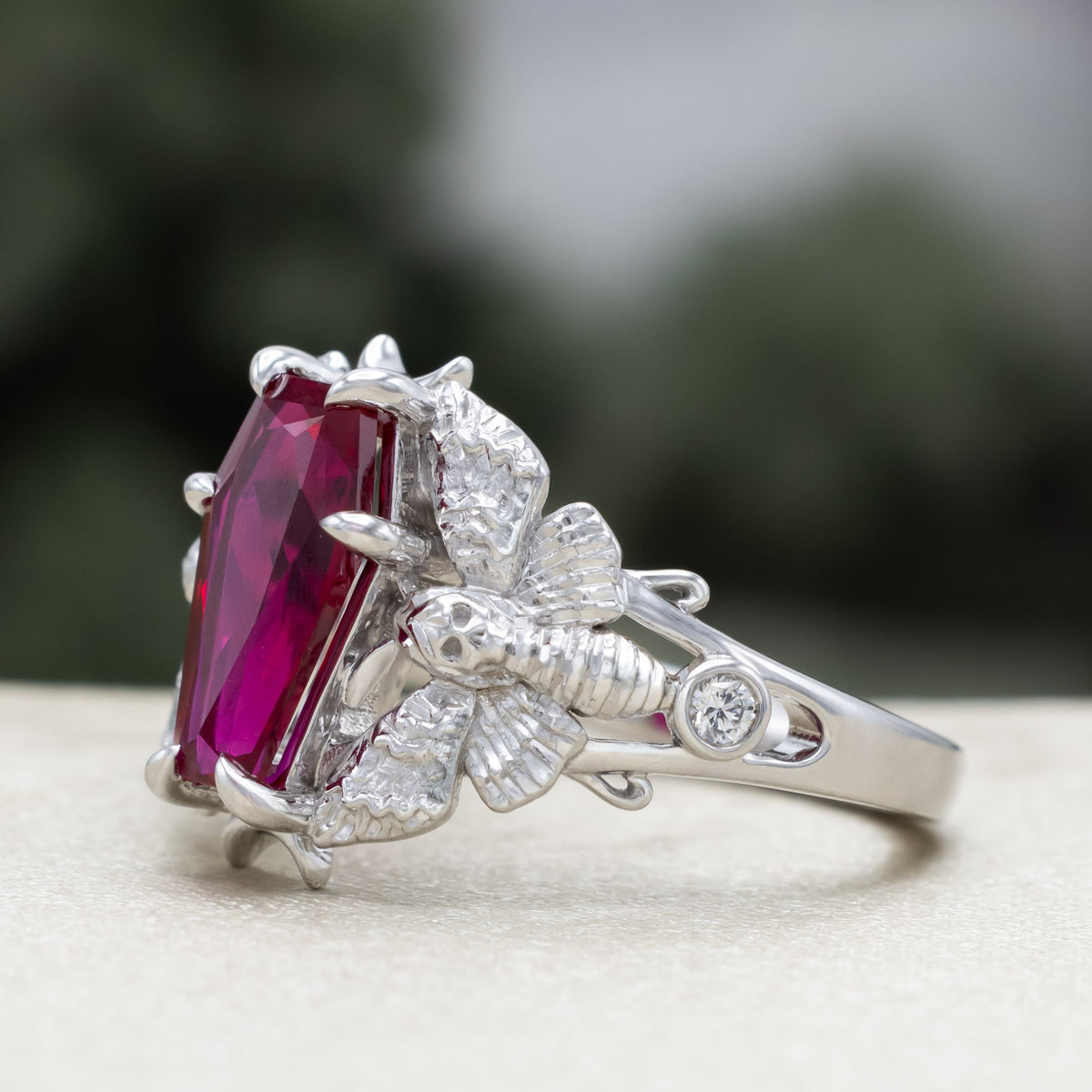 White gold engagement ring with deadhead moth filigree, coffin cut ruby center stone, and diamond accents against a blurred green background