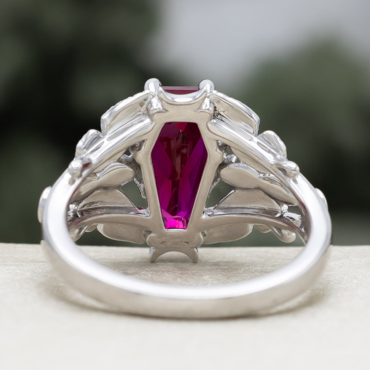 White gold engagement ring with deadhead moth filigree, coffin cut ruby center stone, and diamond accents against a blurred green background