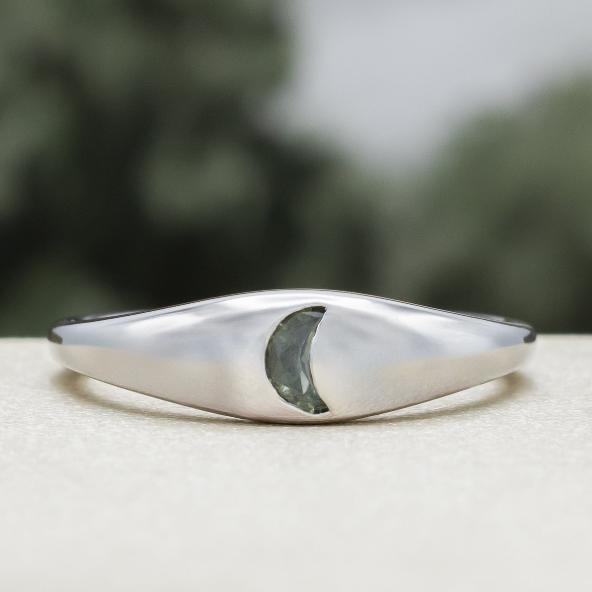 White gold ring with crescent moon shaped sapphire against a blurred green background