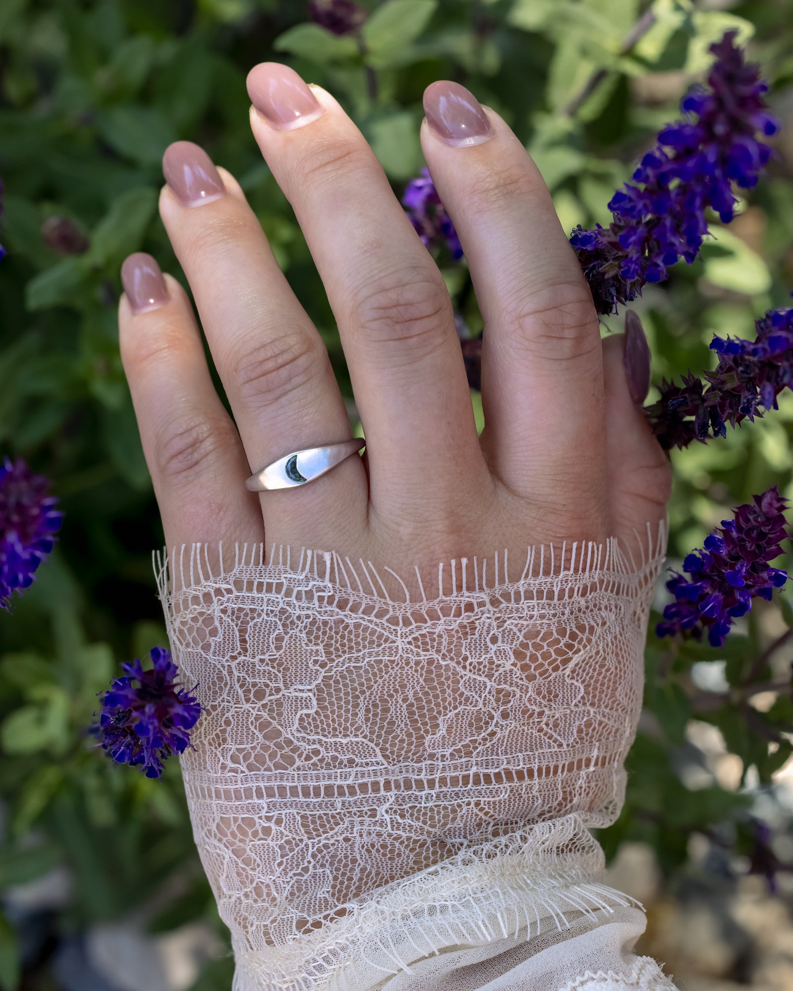 Hand wearing a white gold ring with a crescent moon shaped sapphire against a blurred floral background