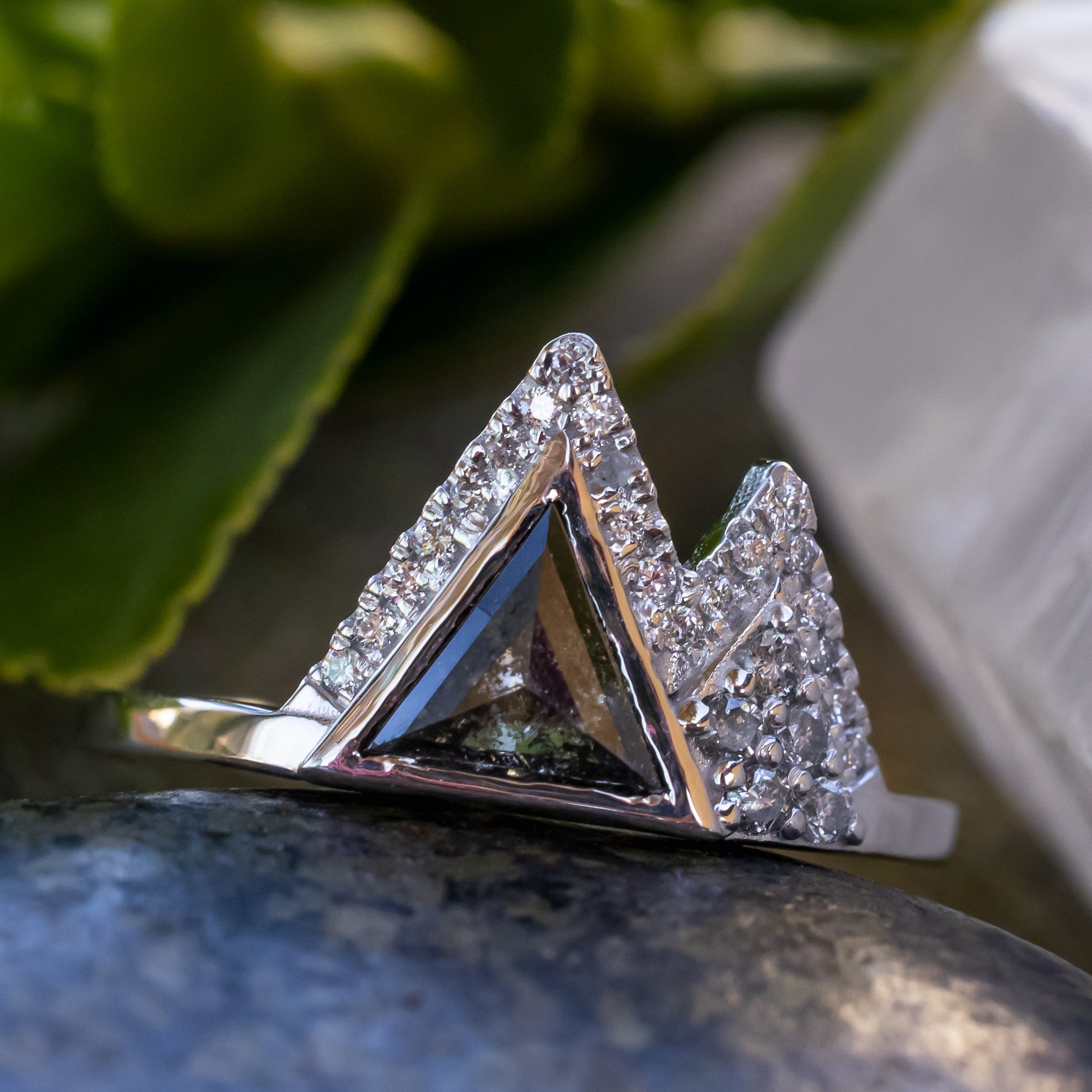 White gold ring with salt & pepper diamond center stone and diamond accents on a stone surface with greenery in the background