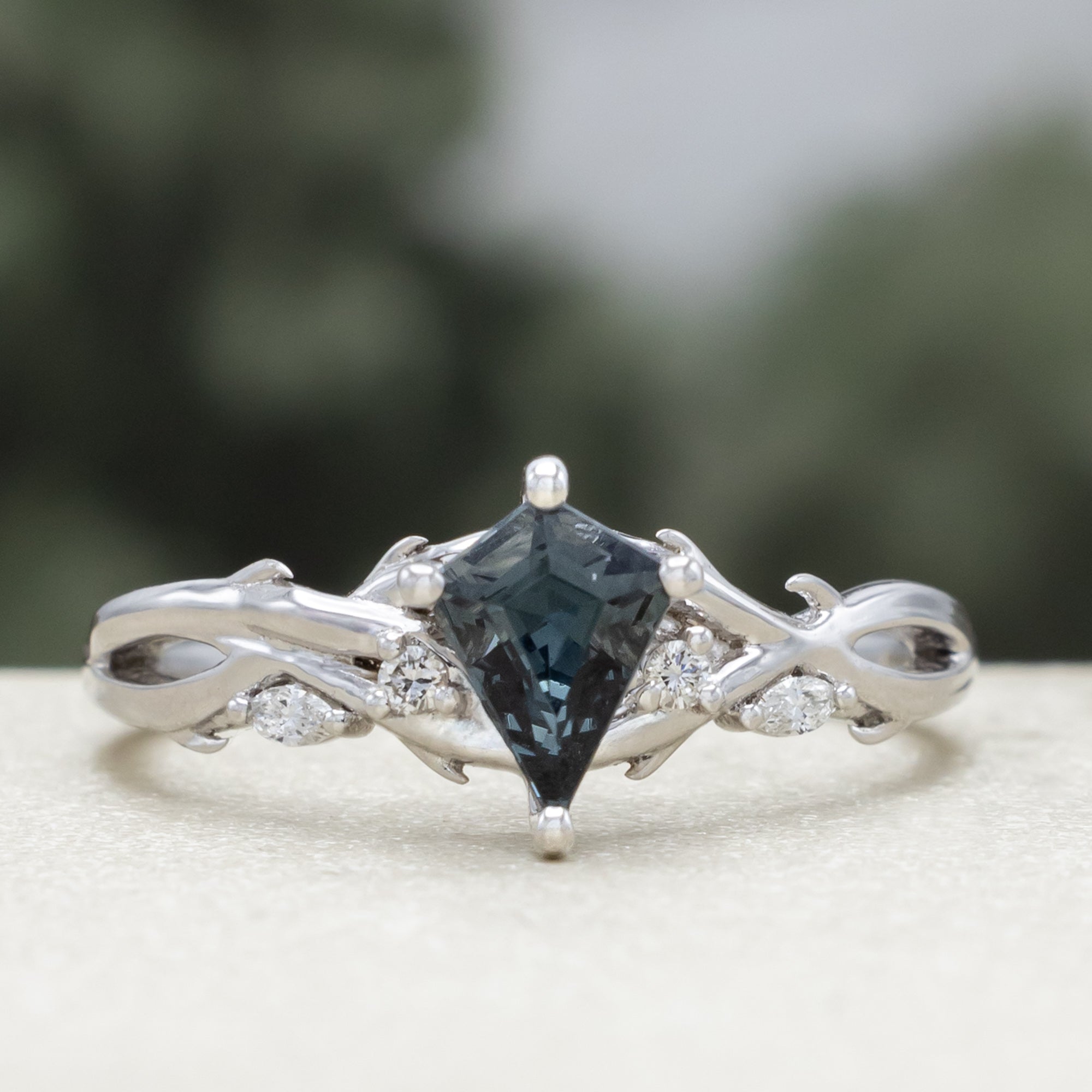 White gold ring with blue spinel center stone & diamond accents against a blurred green background