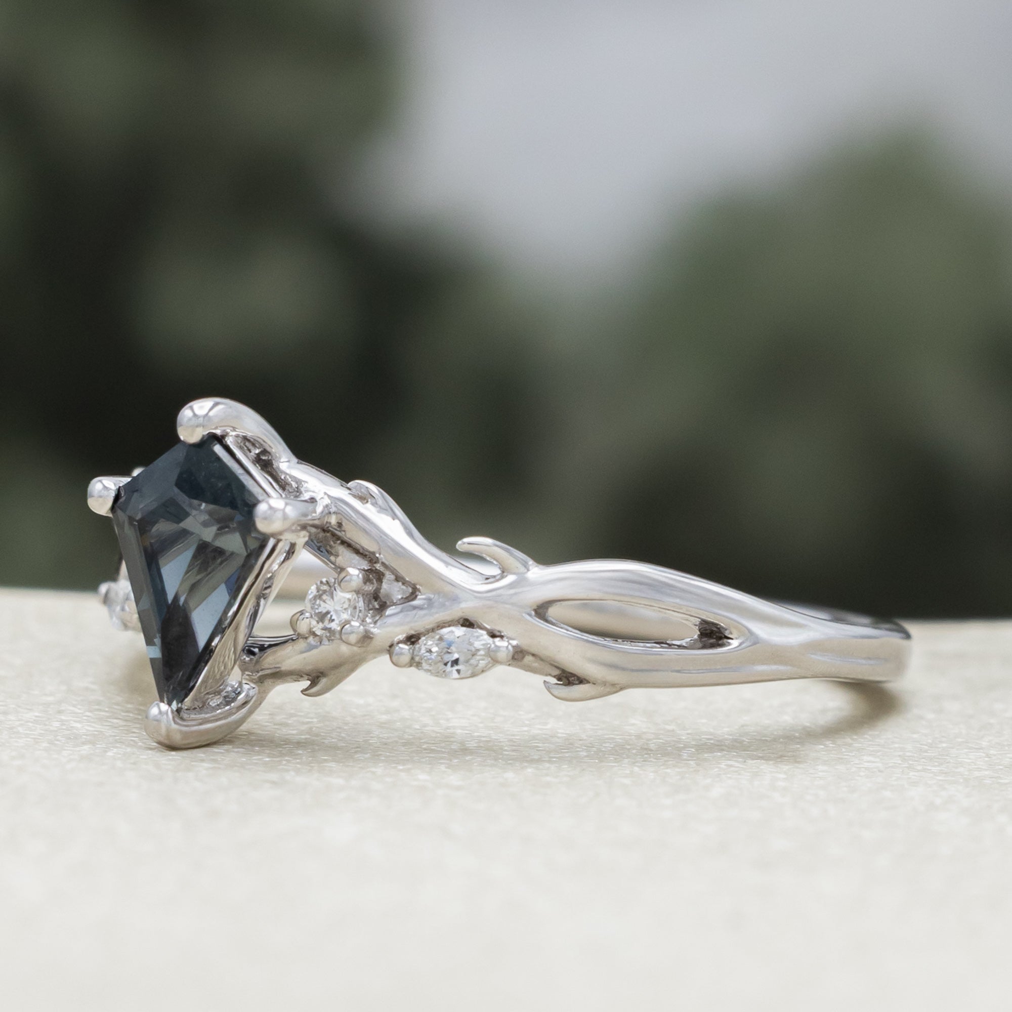 White gold ring with blue spinel center stone & diamond accents against a blurred green background