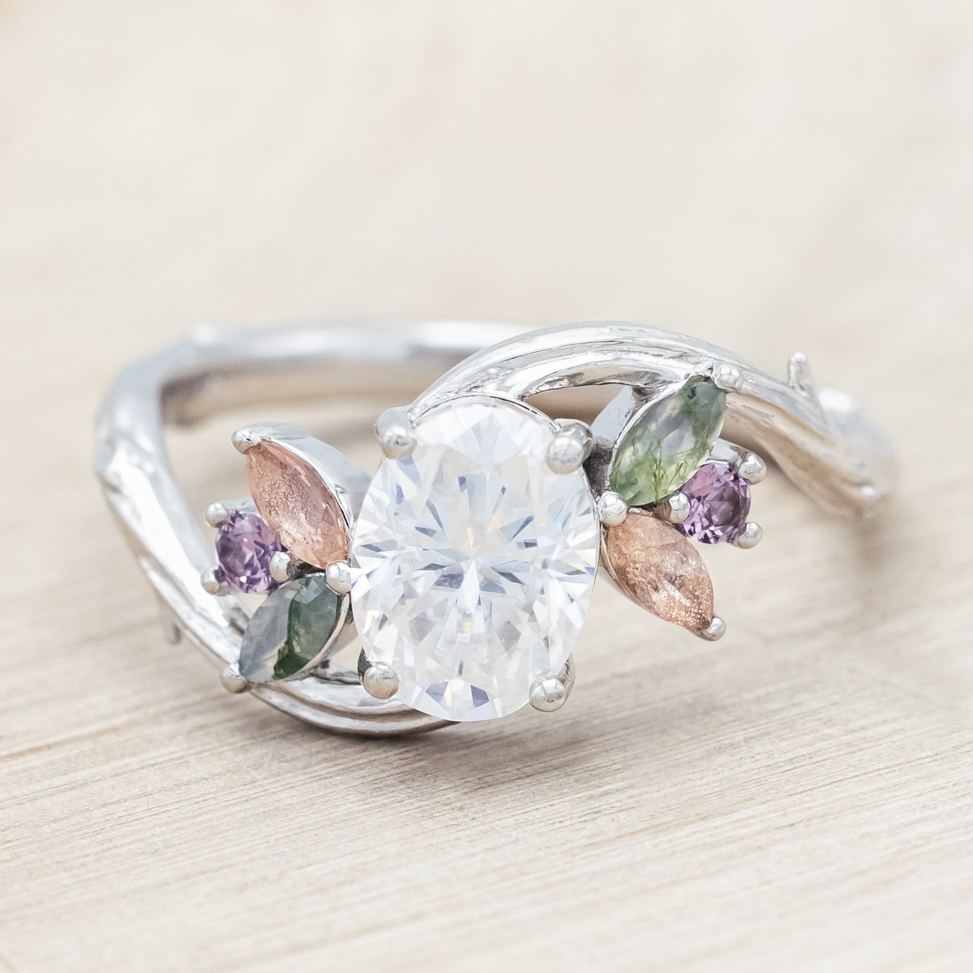 White gold ring with moissanite and smaller spinel, moss agate and sunstone accents on a wooden surface