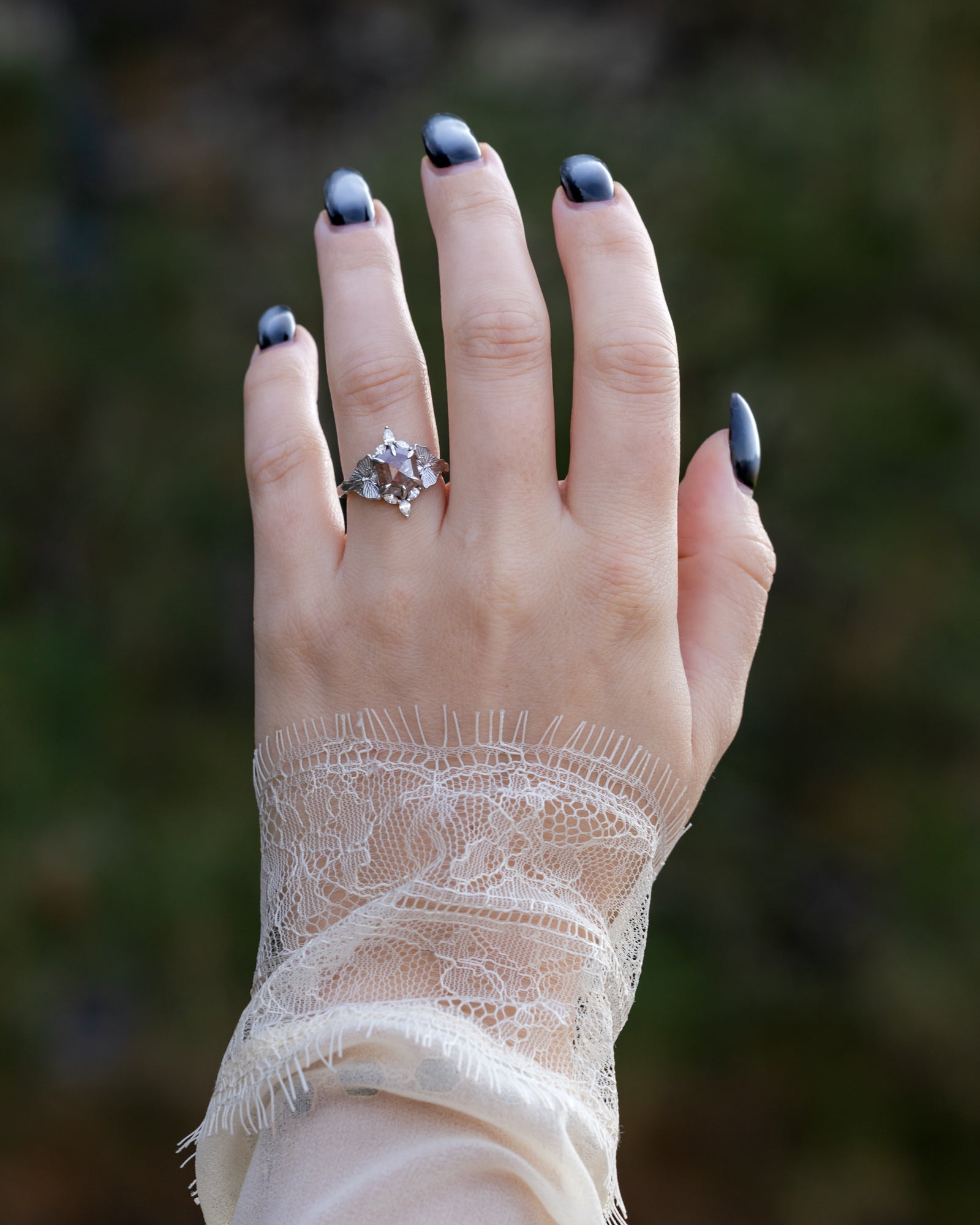 Hand wearing a salt & pepper diamond ring with lace sleeve against a blurred natural background