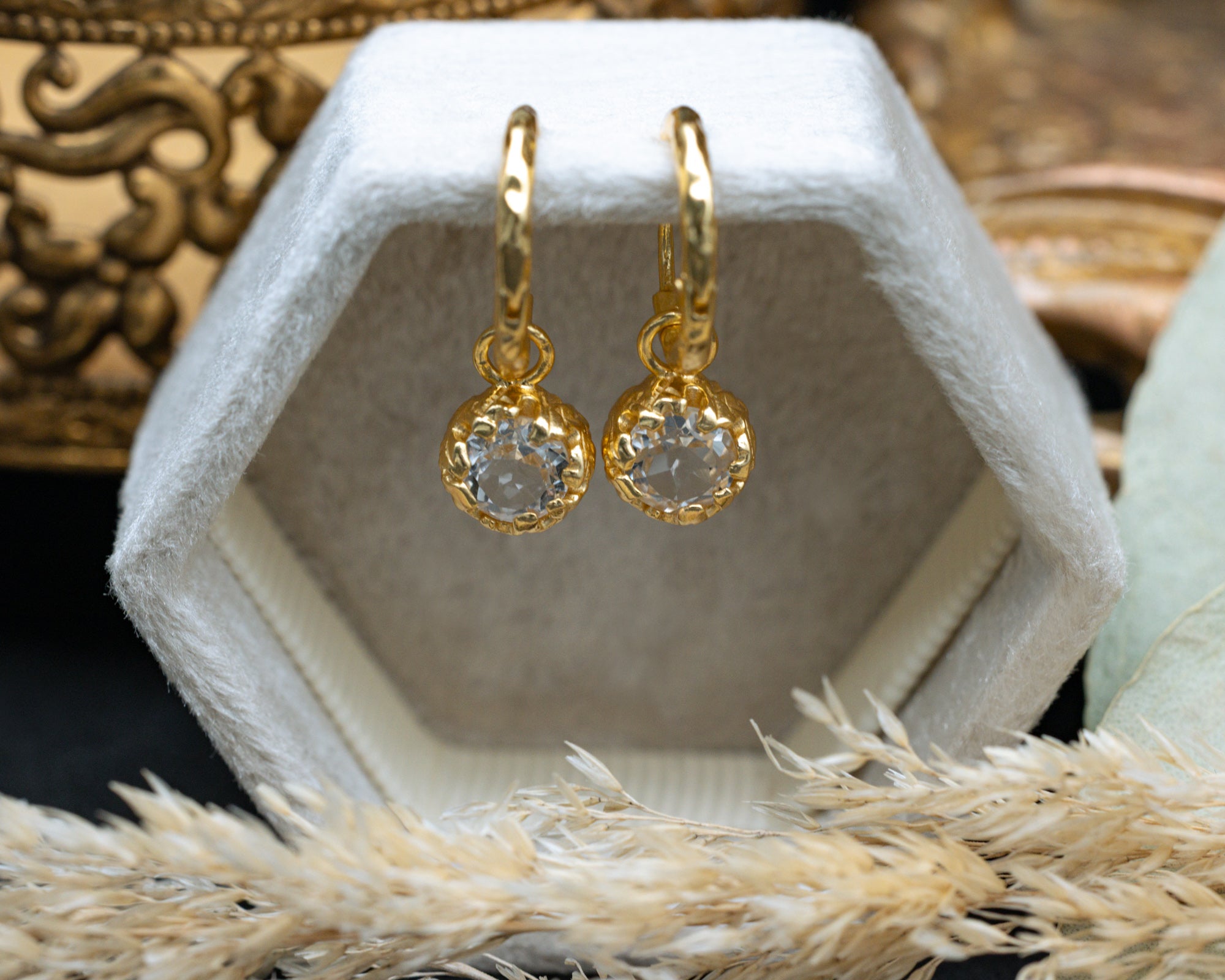 Gold earrings with white topaz in a hexagonal white display box on a textured surface.