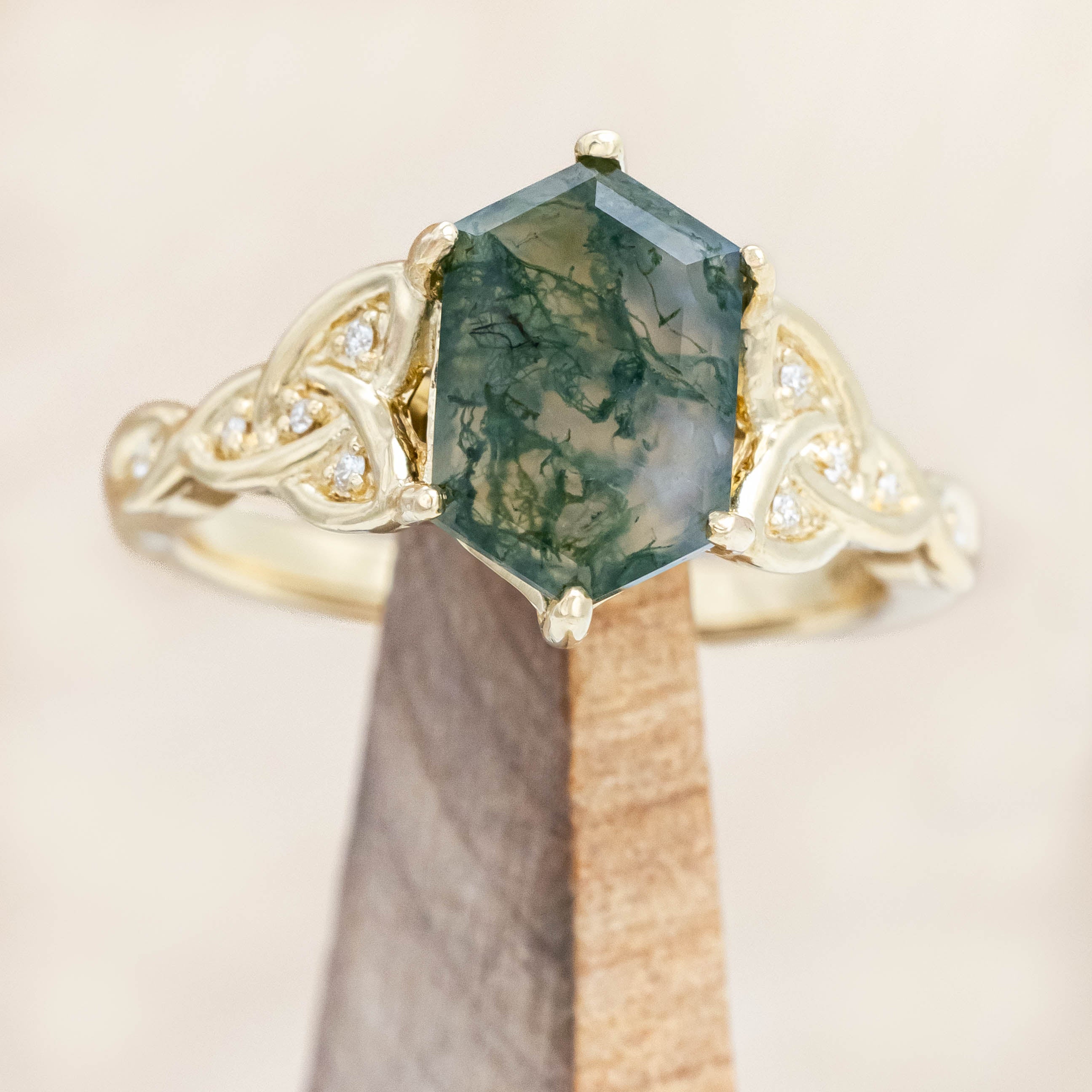 Gold ring with triquetra knot accent and moss agate on a wooden stand against a beige background
