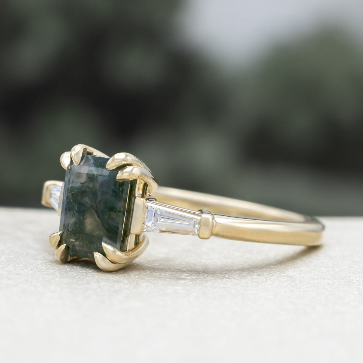Gold ring with emerald cut moss agate center stone & diamond accents against a blurred green background