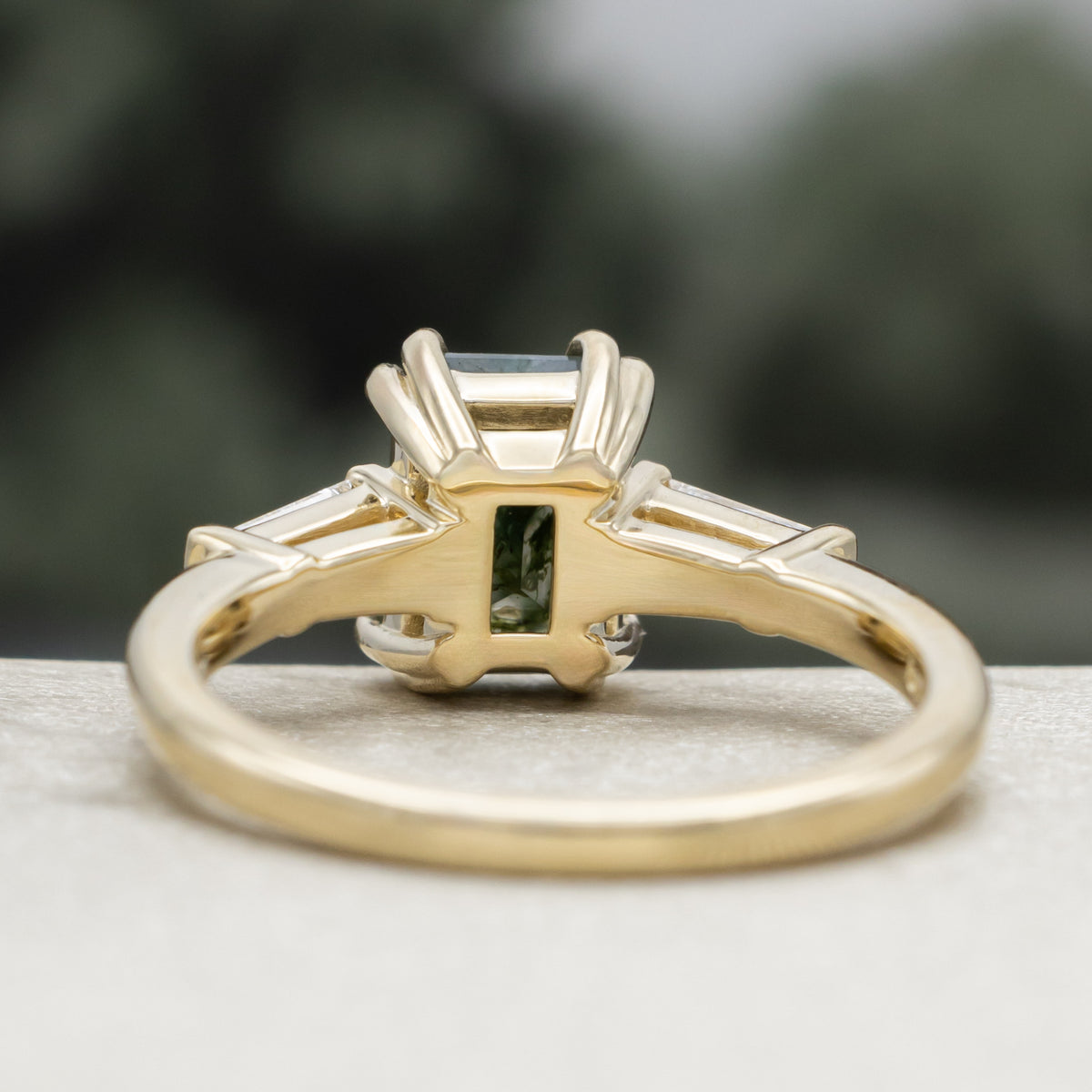 Gold ring with emerald cut moss agate center stone & diamond accents against a blurred green background