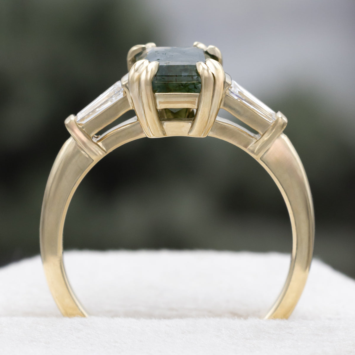 Gold ring with emerald cut moss agate center stone & diamond accents against a blurred green background