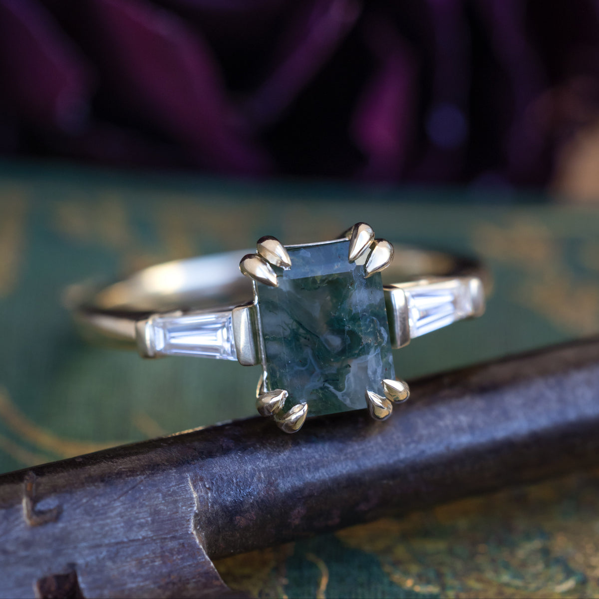 Gold ring with emerald cut moss agate center stone & diamond accents against a blurred, ornate background