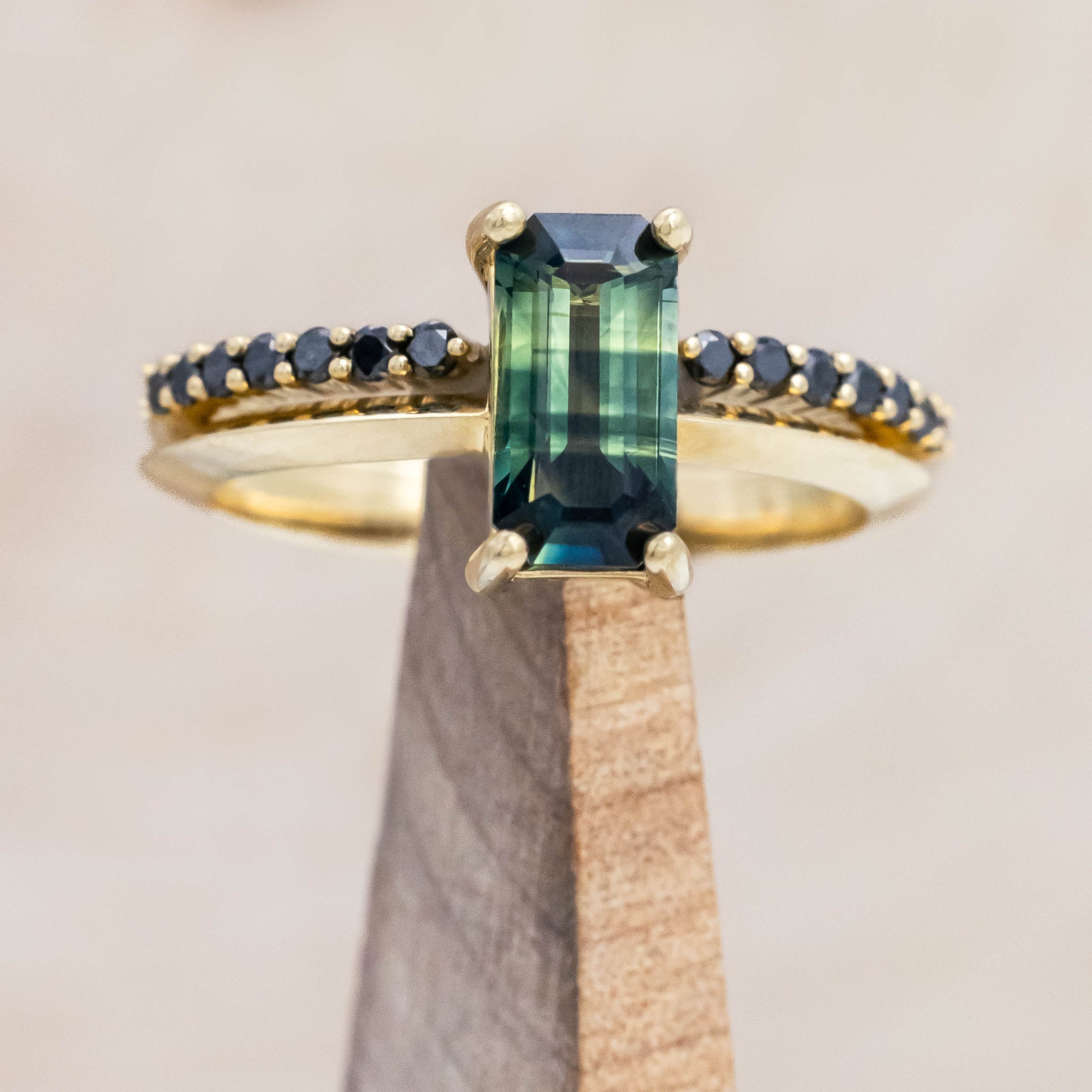 14K yellow gold ring with a green sapphire and black diamond tracer on a wooden stand against a beige background