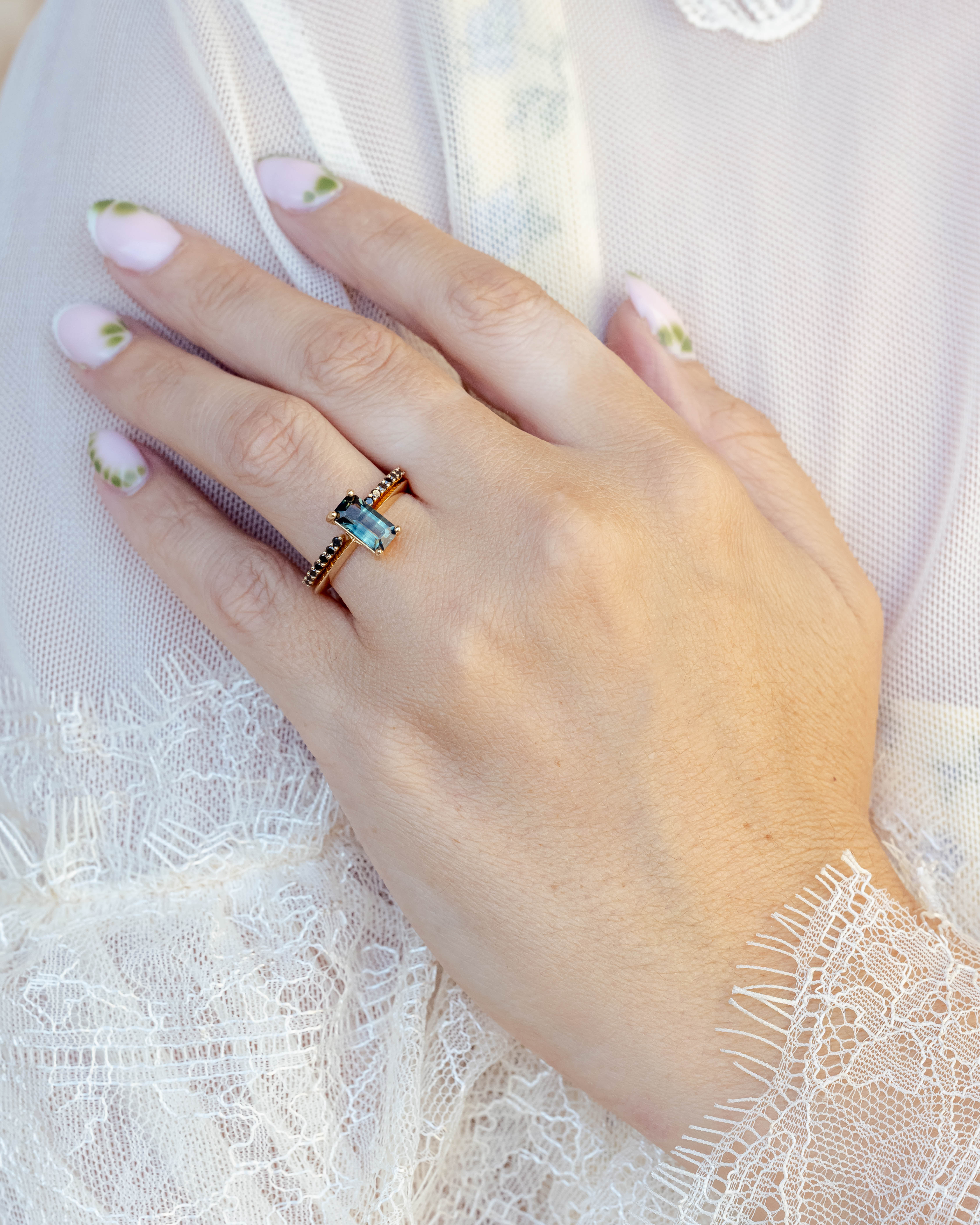 Hand wearing a ring with a green sapphire and black diamond tracer on a white lace fabric background
