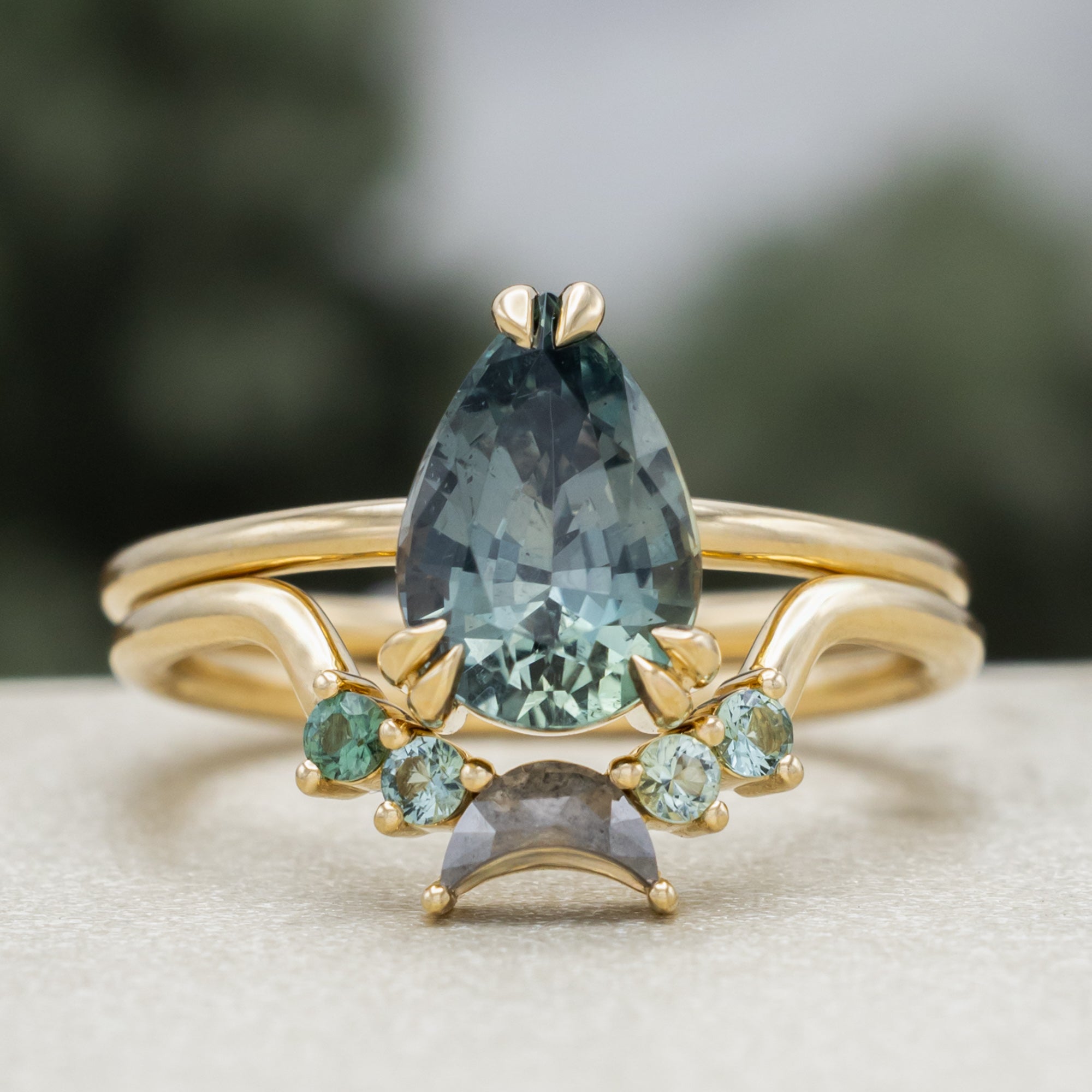 A gold ring featuring a pear-cut blue sapphire with tourmaline and salt & pepper diamond tracer on a blurred green background.