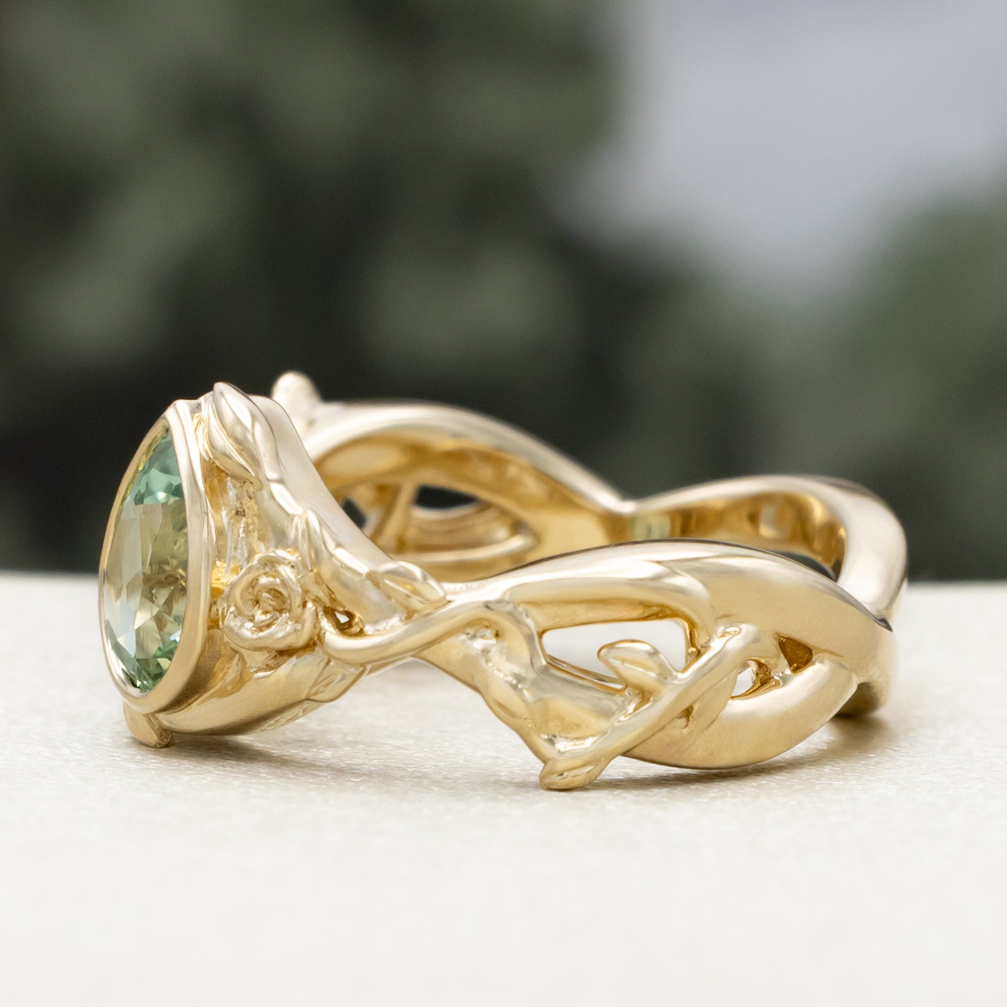 Gold ring with pear lab sapphire and rose/vine accents on a blurred background
