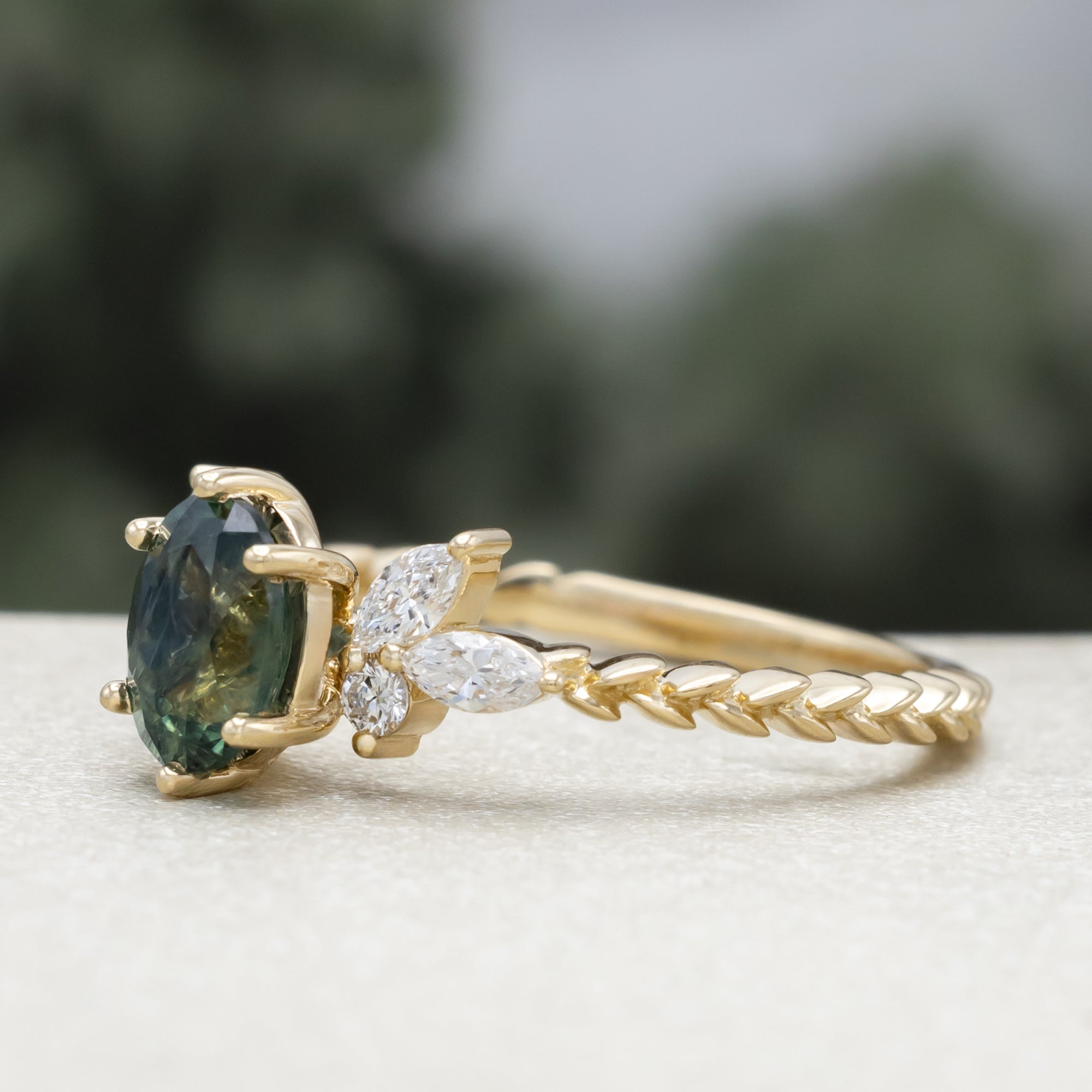 A yellow gold ring featuring an oval-cut Montana sapphire with diamond accents on a vine band against a blurred green background.