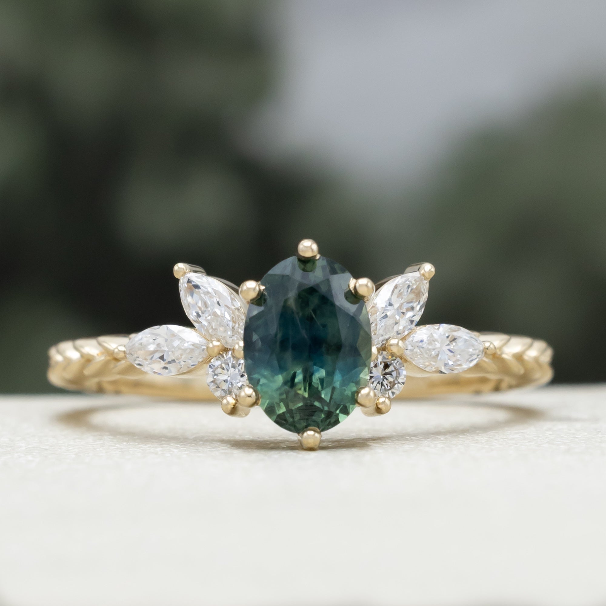 A yellow gold ring featuring an oval-cut Montana sapphire with diamond accents on a vine band against a blurred green background.