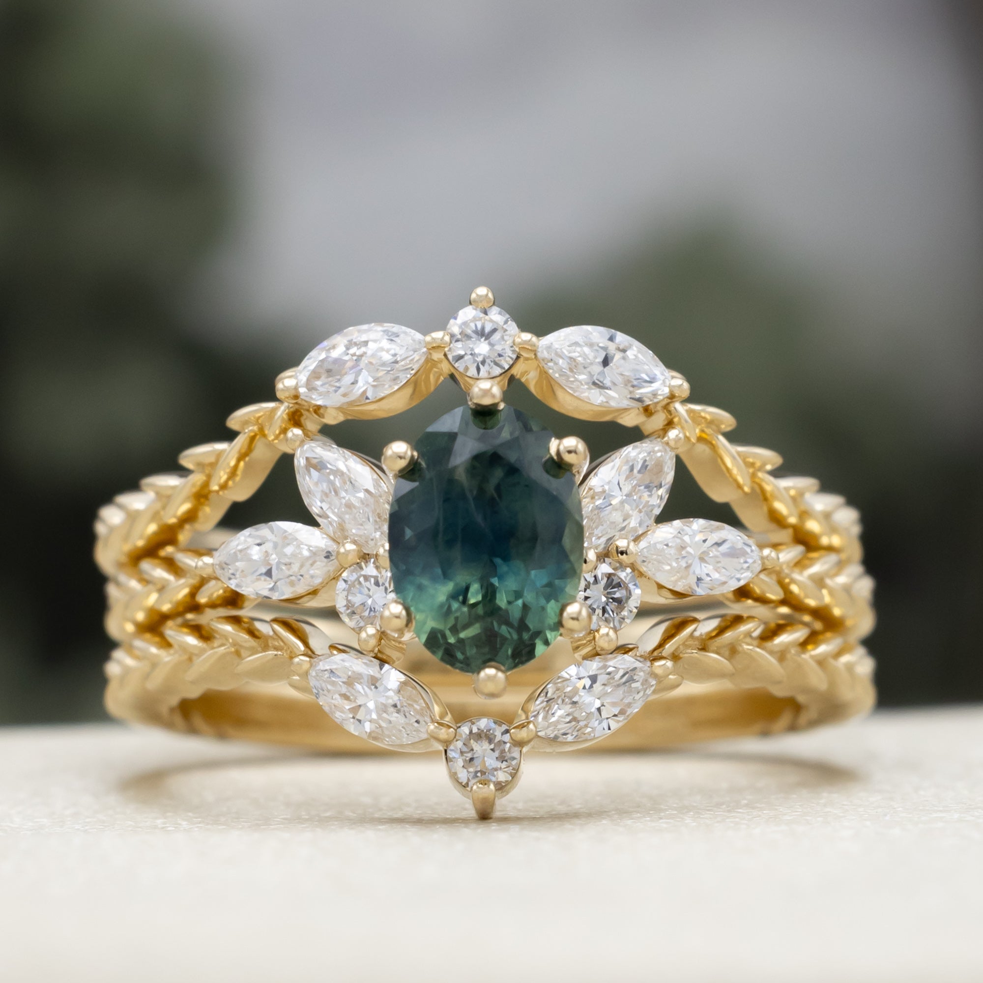 A yellow gold ring featuring an oval-cut Montana sapphire with diamond accents and tracers on a vine band against a blurred green background.