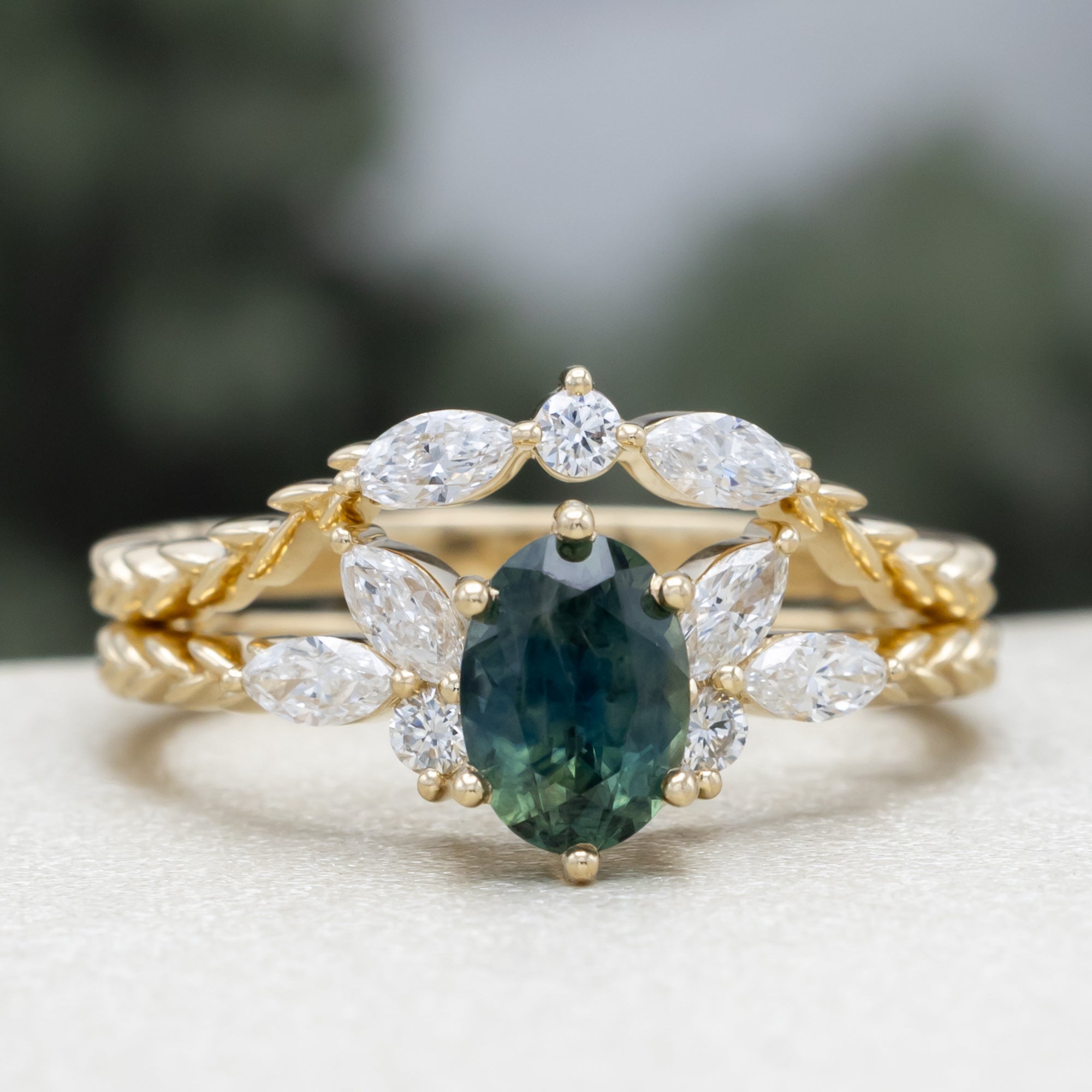 A yellow gold ring featuring an oval-cut Montana sapphire with diamond accents and tracer on a vine band against a blurred green background.