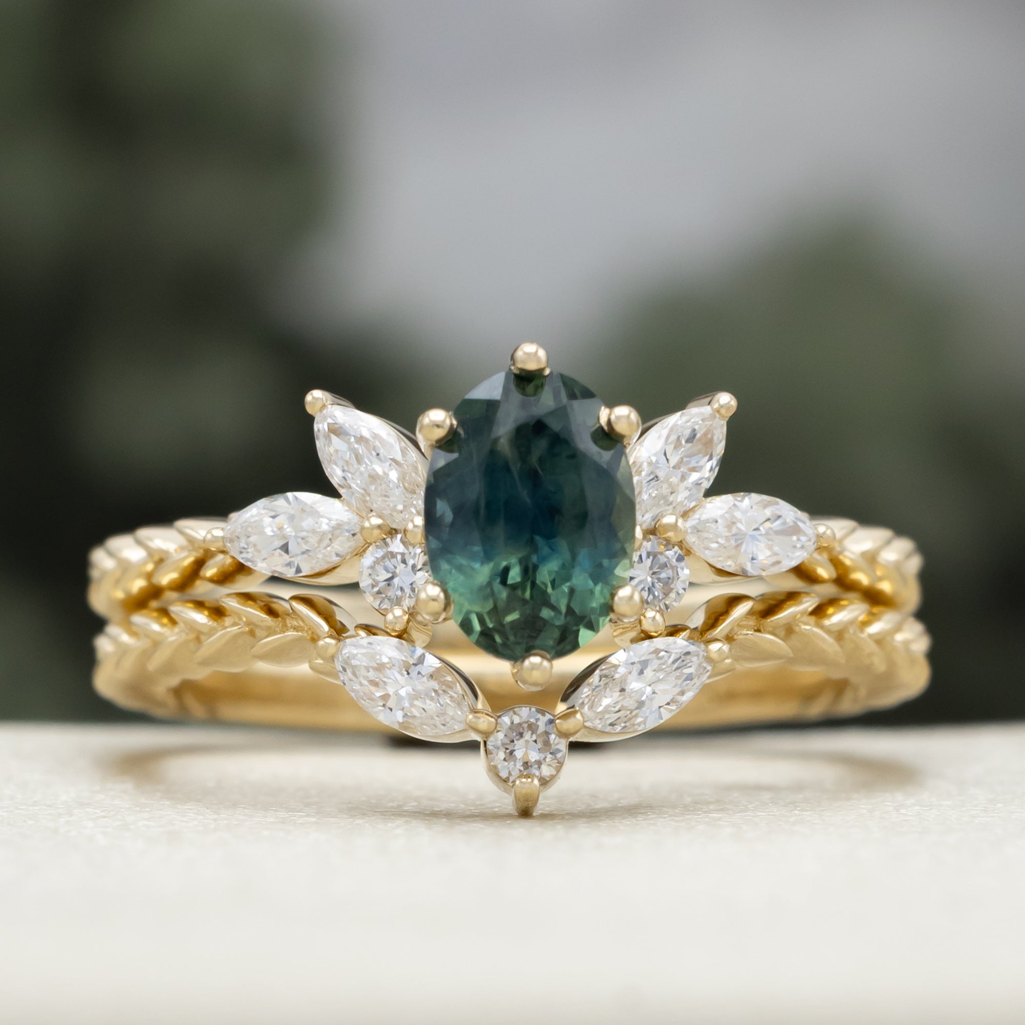 A yellow gold ring featuring an oval-cut Montana sapphire with diamond accents and tracer on a vine band against a blurred green background.
