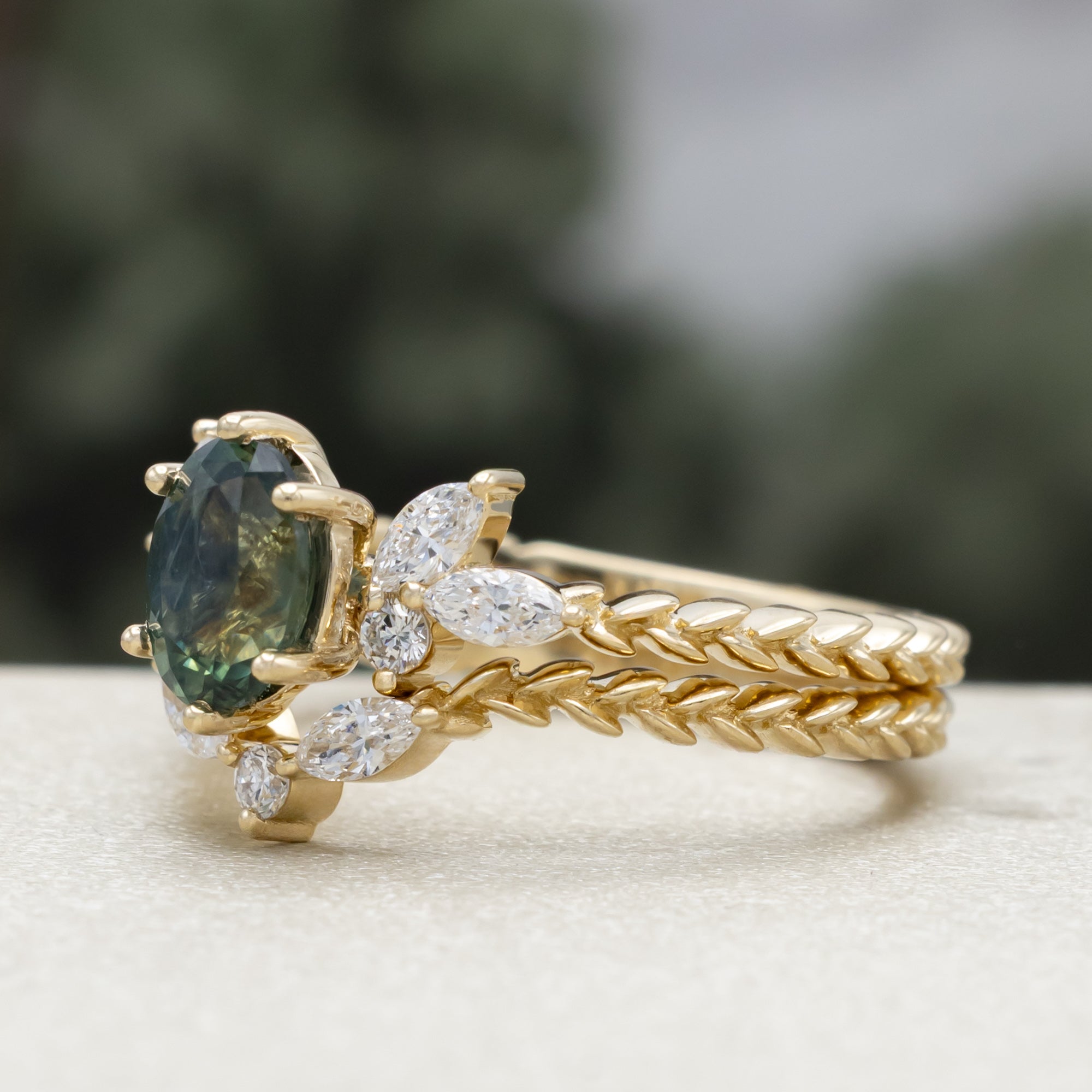 A yellow gold ring featuring an oval-cut Montana sapphire with diamond accents and tracer on a vine band against a blurred green background.