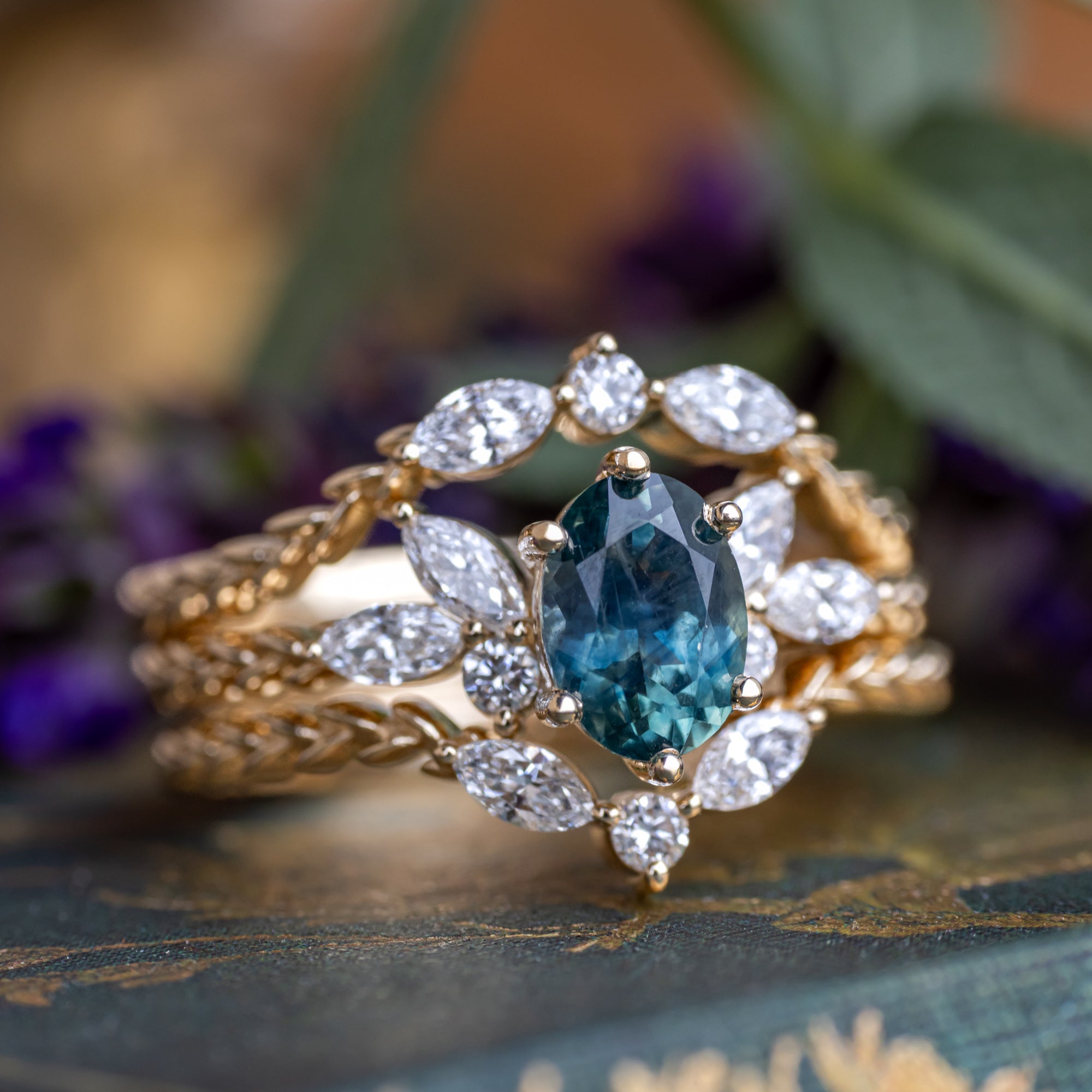 A yellow gold ring featuring an oval-cut Montana sapphire with diamond accents and tracers on a decorative surface with blurred flowers in the background
