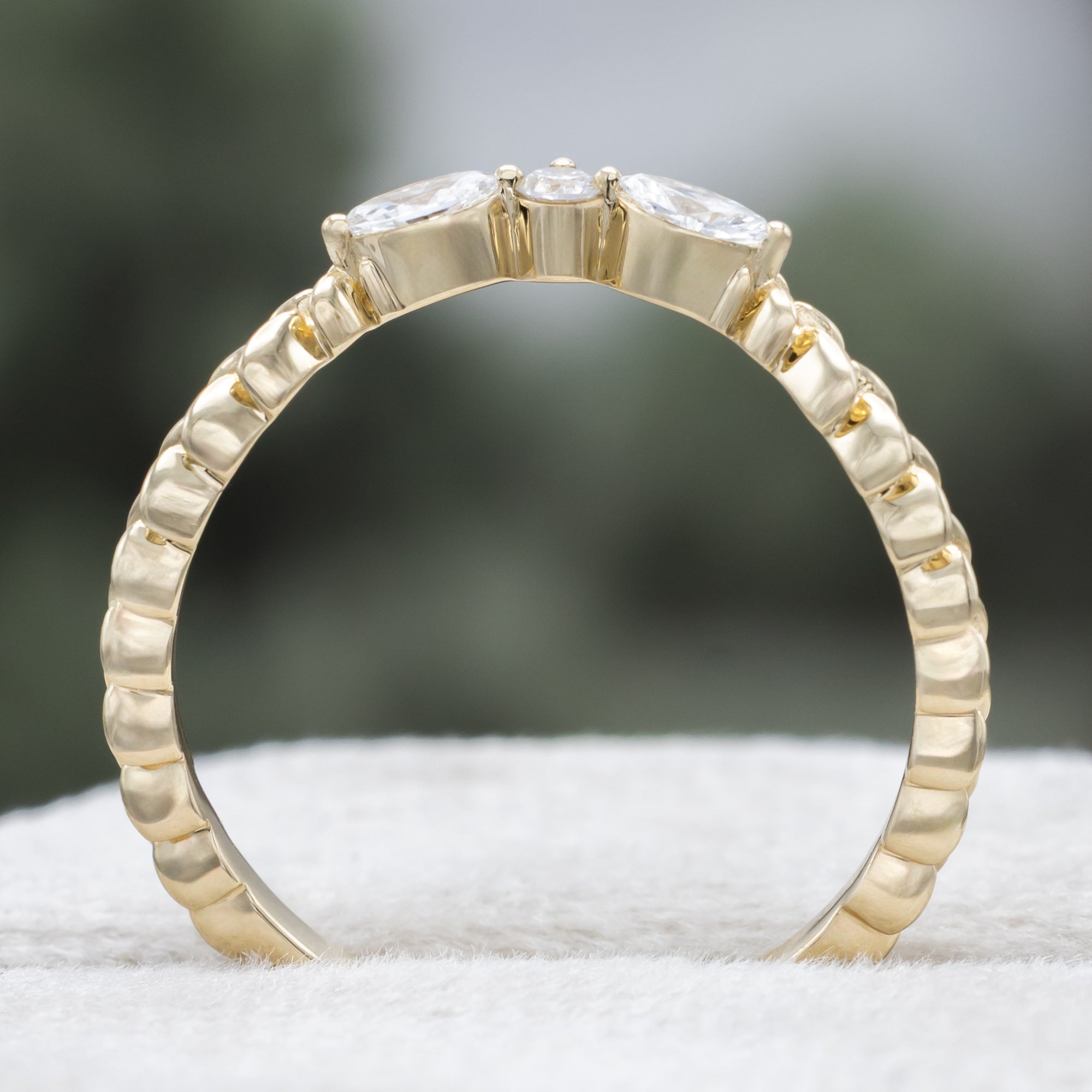 A yellow gold ring tracer with three diamond accents on a vine style band against a blurred green background
