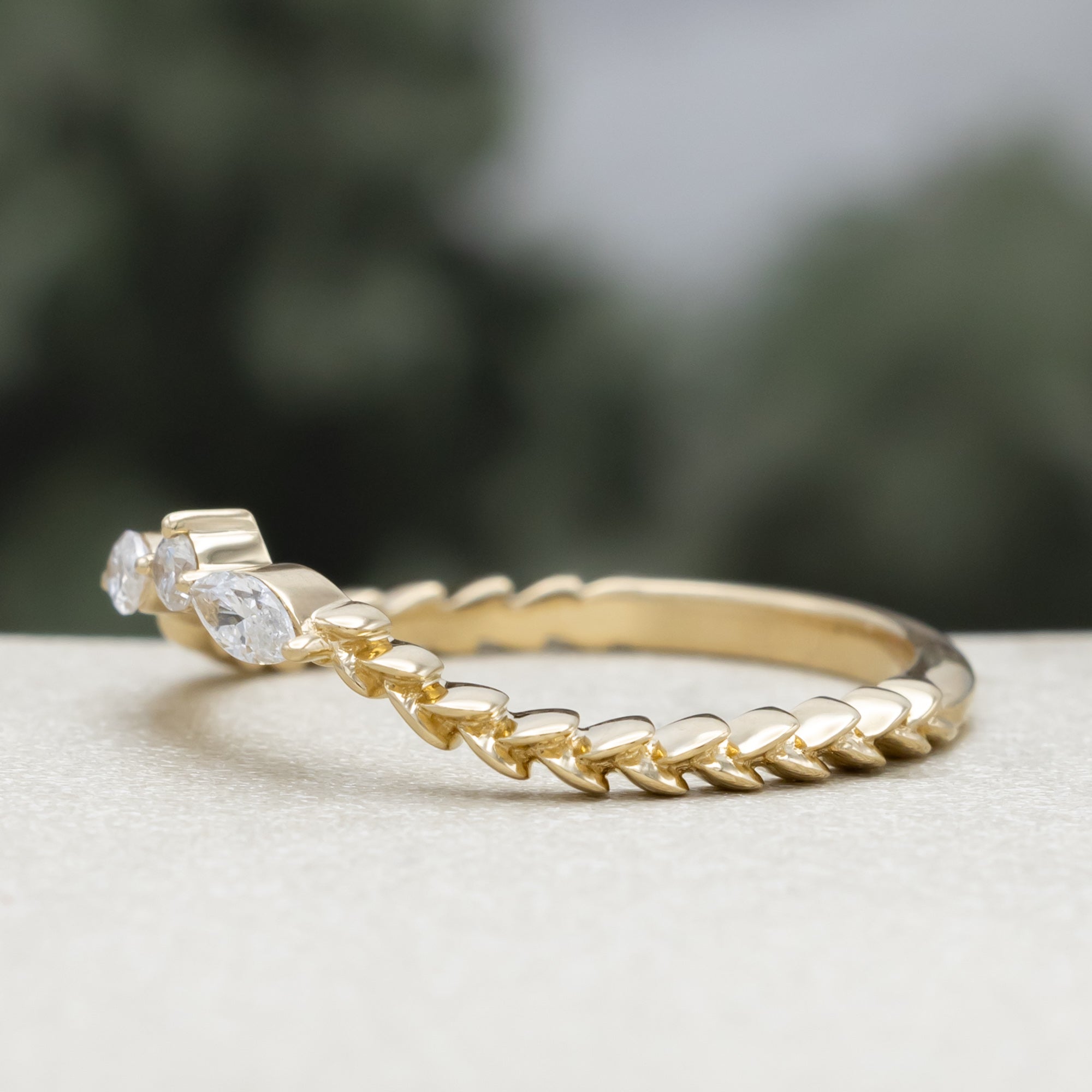 A yellow gold ring tracer with three diamond accents on a vine style band against a blurred green background
