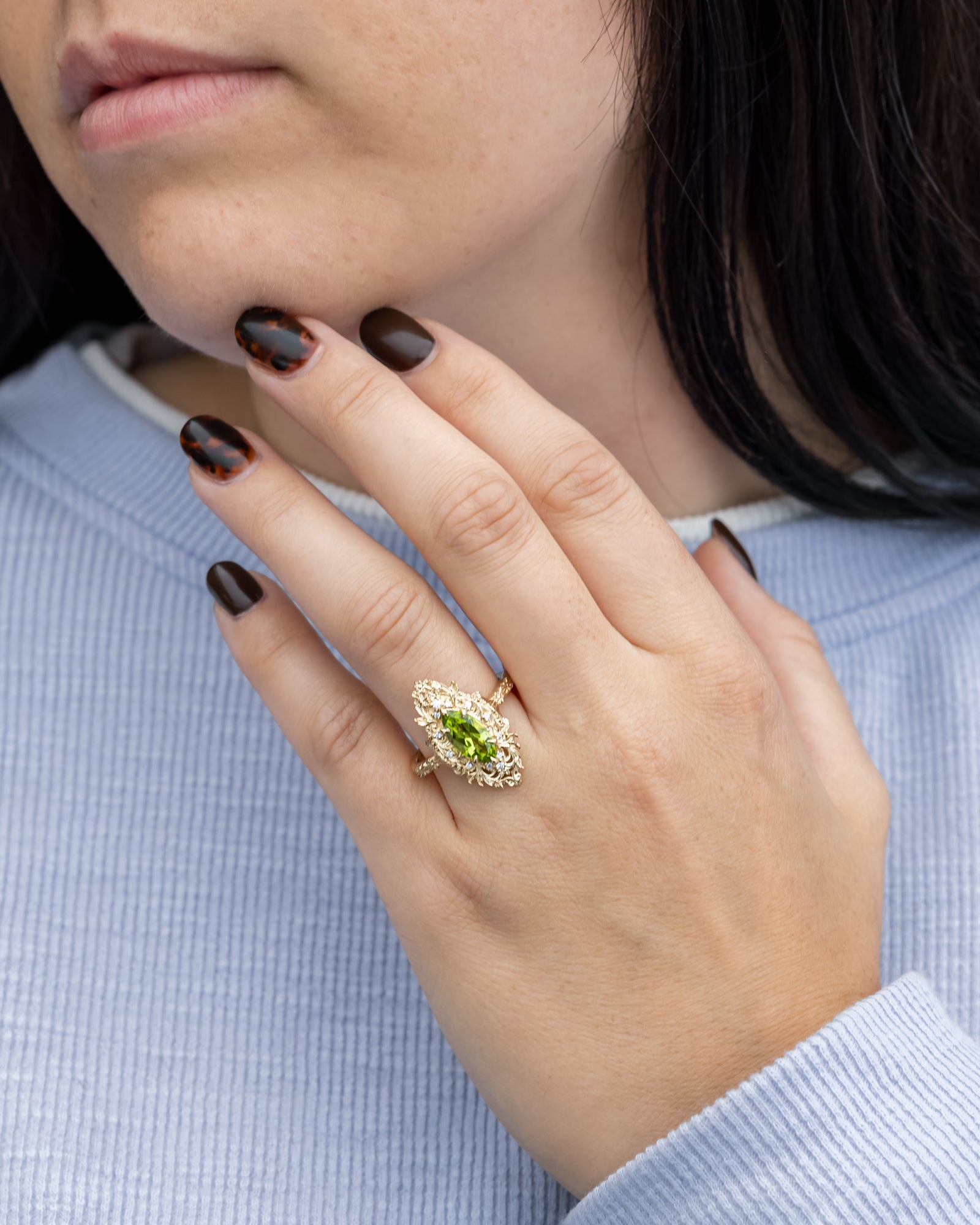 Close-up of a hand wearing an ornate ring with a green peridot, against a light blue background.