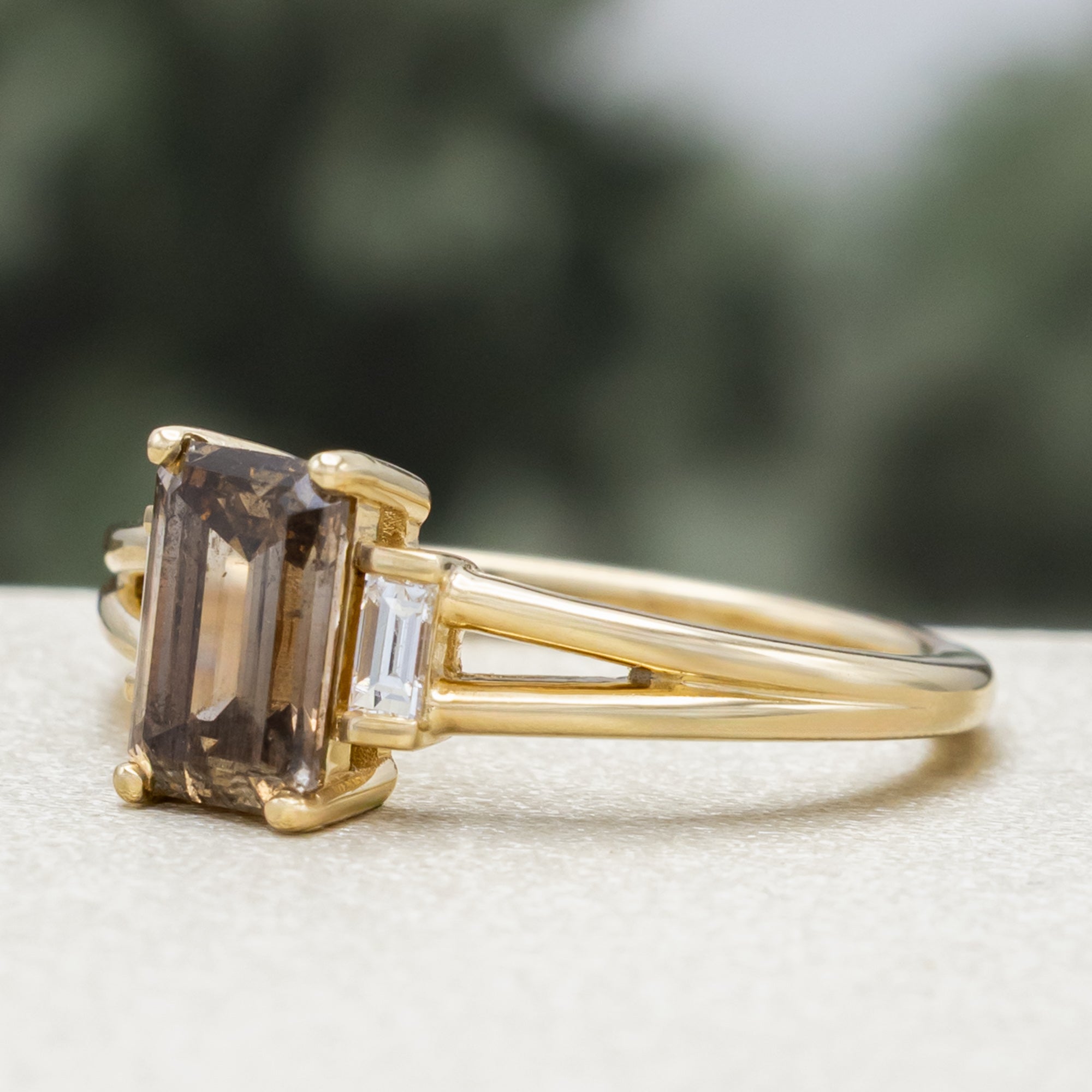 Gold ring with champagne diamond centerstone & diamond accent against a blurred green background
