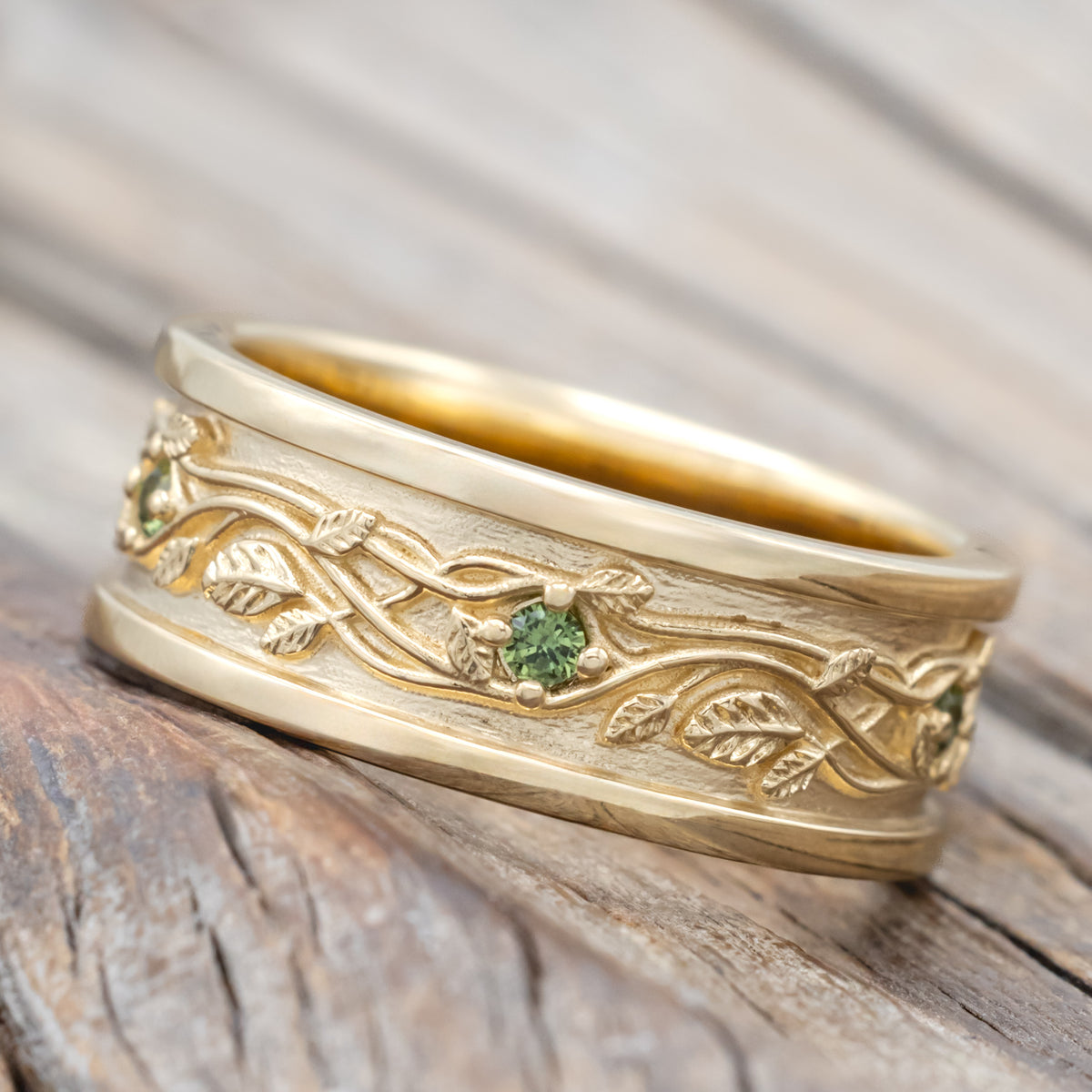 Yellow gold band with vine filigree and green sapphire accents against a blurred wood background