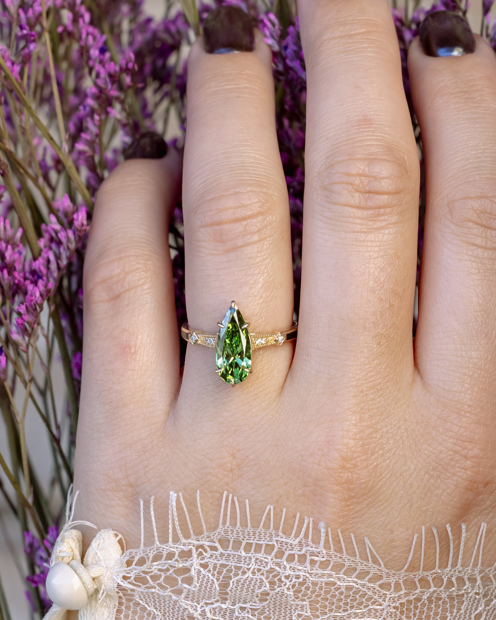 Hand wearing a gold ring with a green diamond against a blurred background of purple flowers.