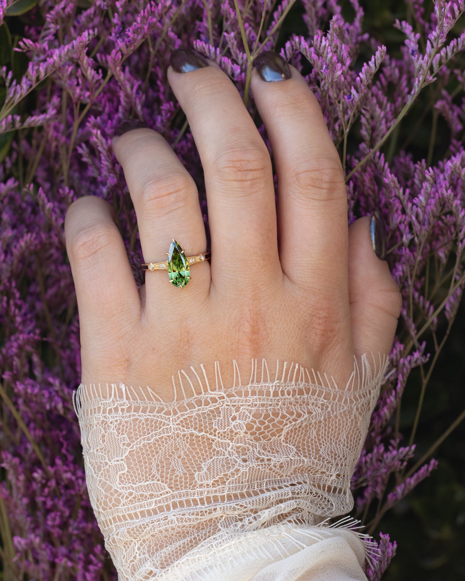 Hand wearing a gold ring with a green diamond against a blurred background of purple flowers.