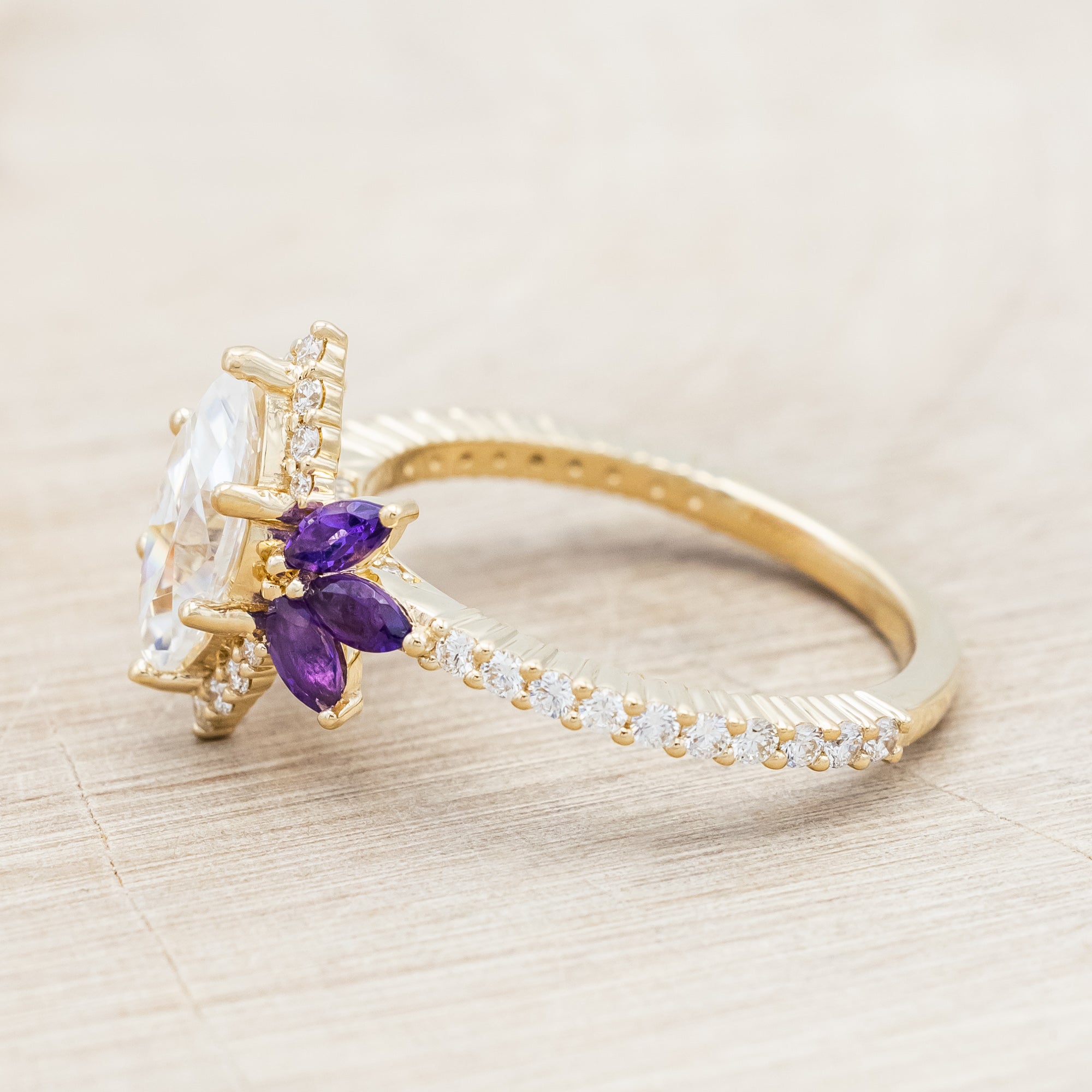 Yellow gold ring with a moissanite center stone and small purple amethyst accents on a beige background