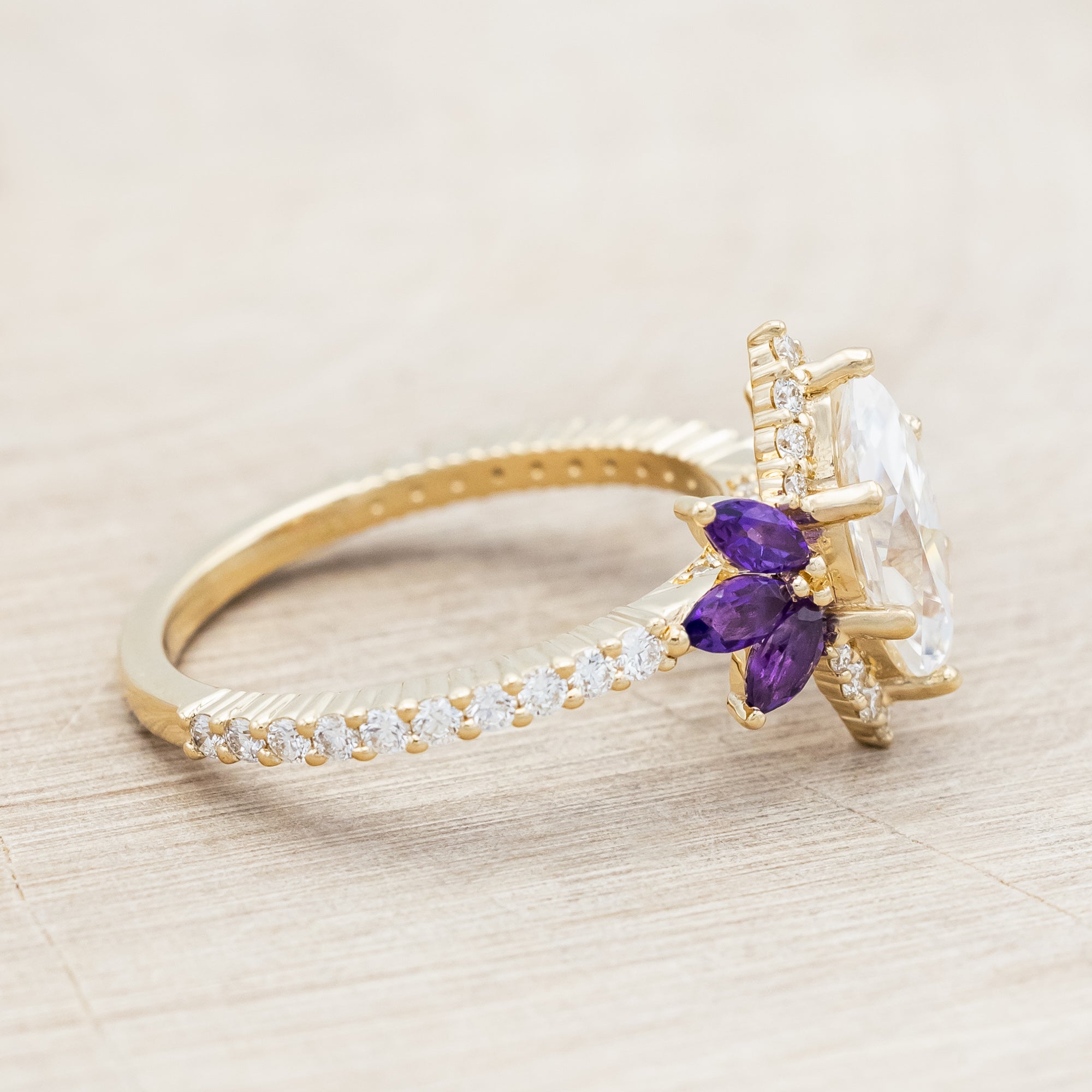 Yellow gold ring with a moissanite center stone and small purple amethyst accents on a beige background