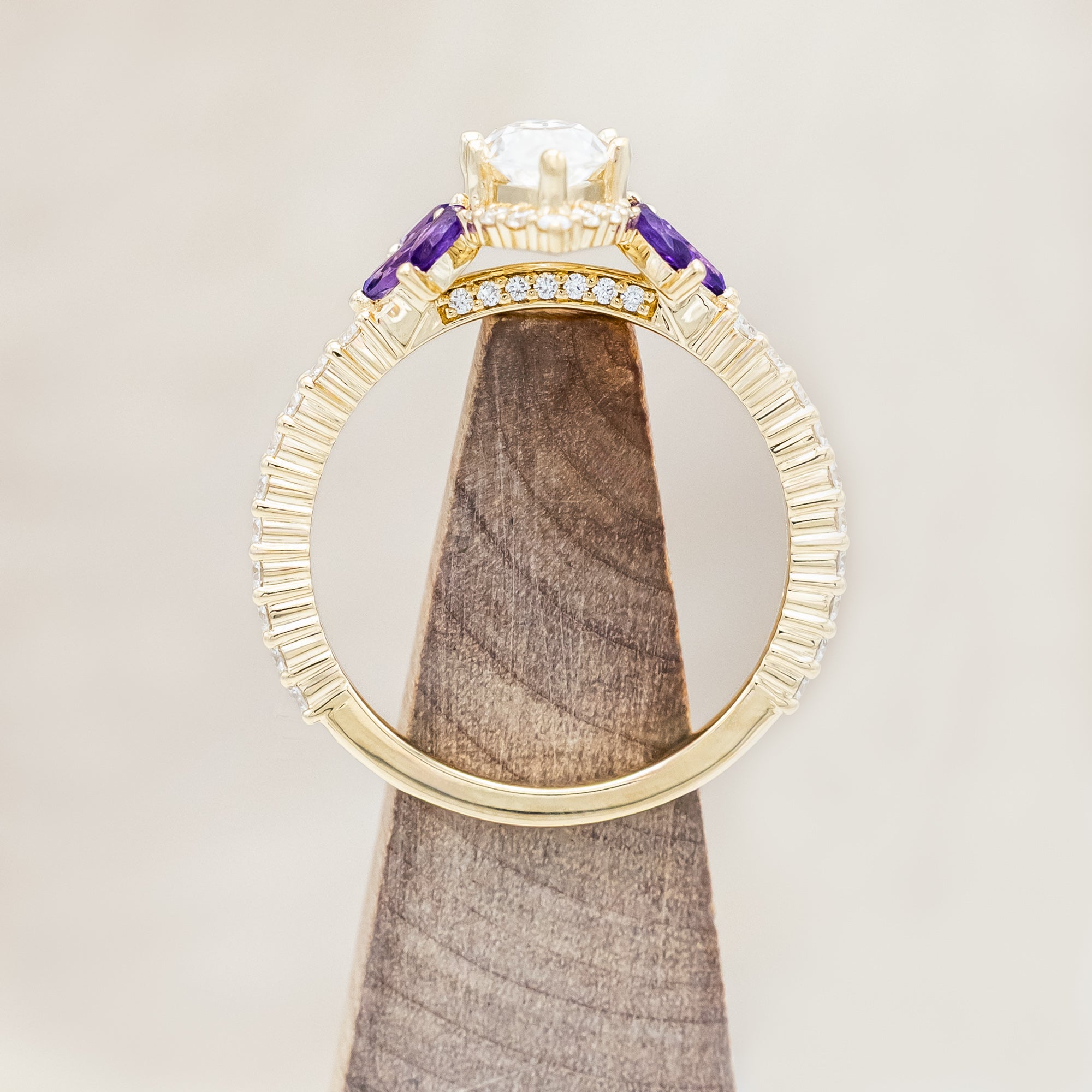 Yellow gold ring with a moissanite center stone and small purple amethyst accents on a beige background