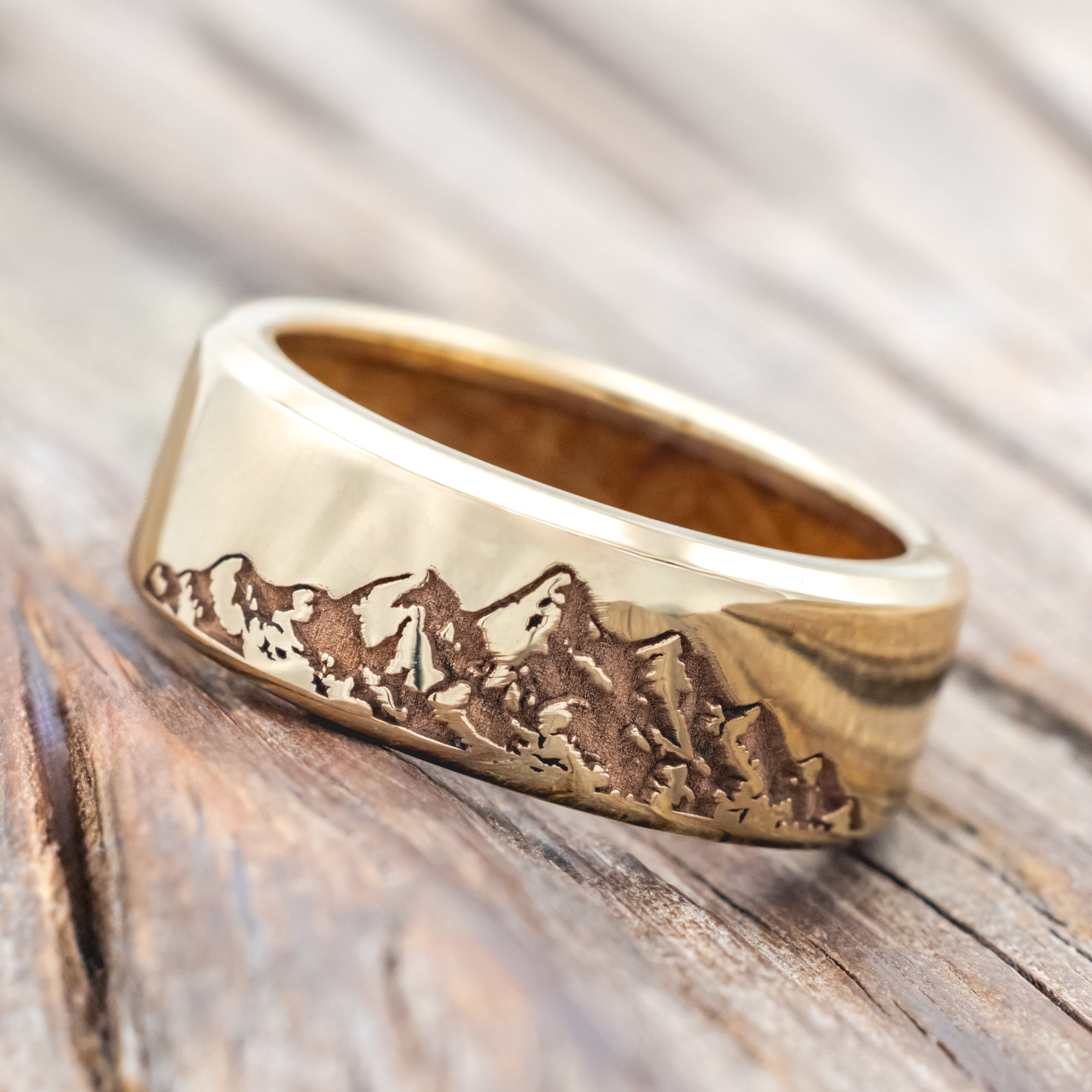 Gold ring with mountain engraving and whiskey barrel lining on a wooden surface
