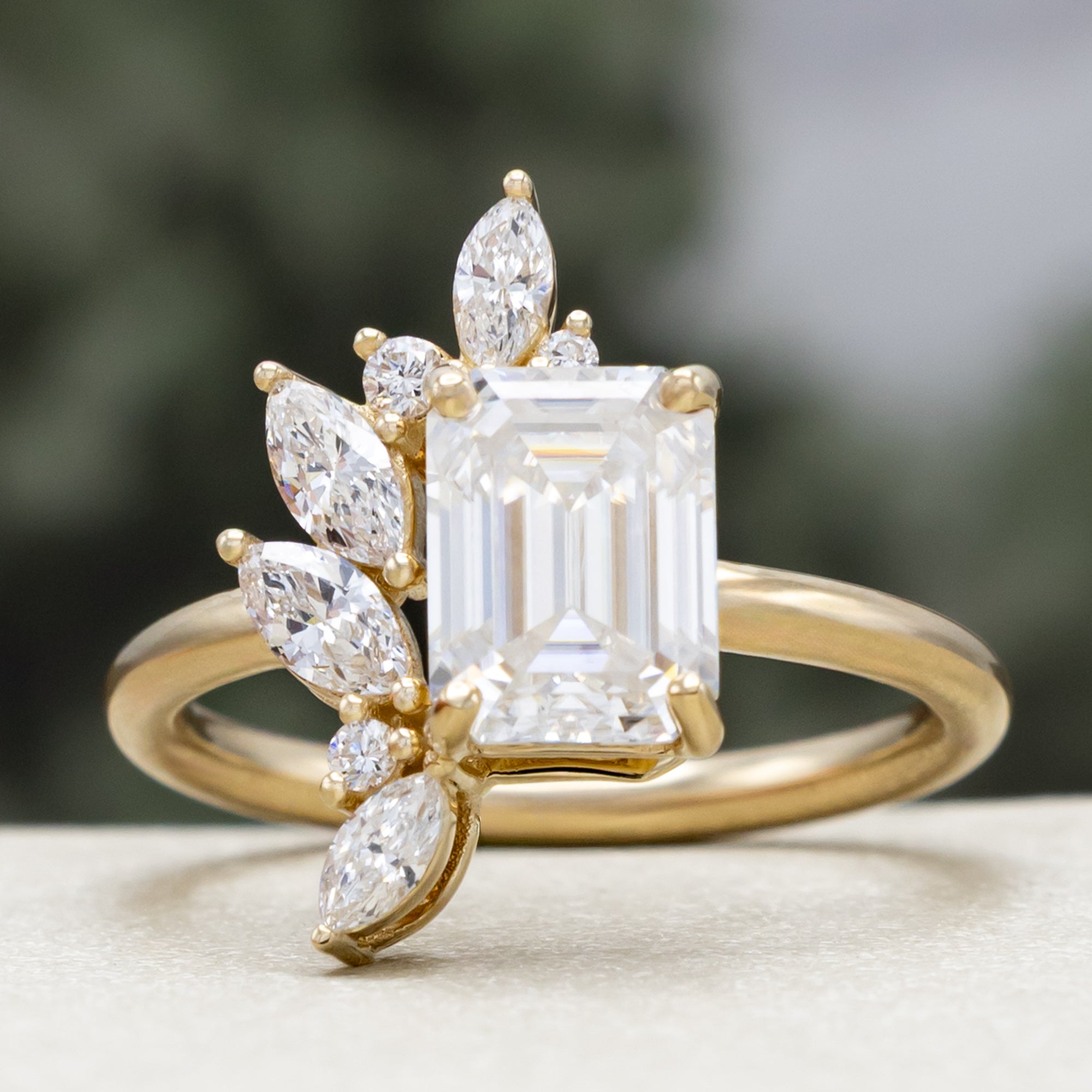 Gold ring with emerald-cut lab diamond and marquise diamond accents on a blurred green background