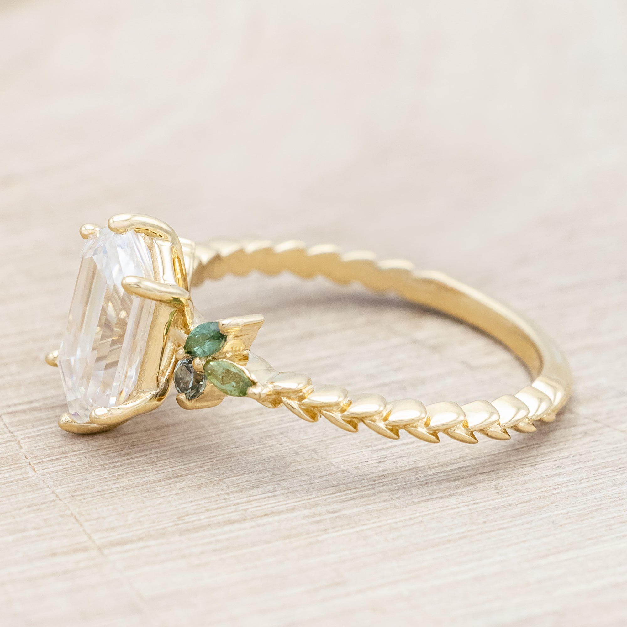 Gold ring with hexagon-cut diamond and green gemstone accents on a beige background